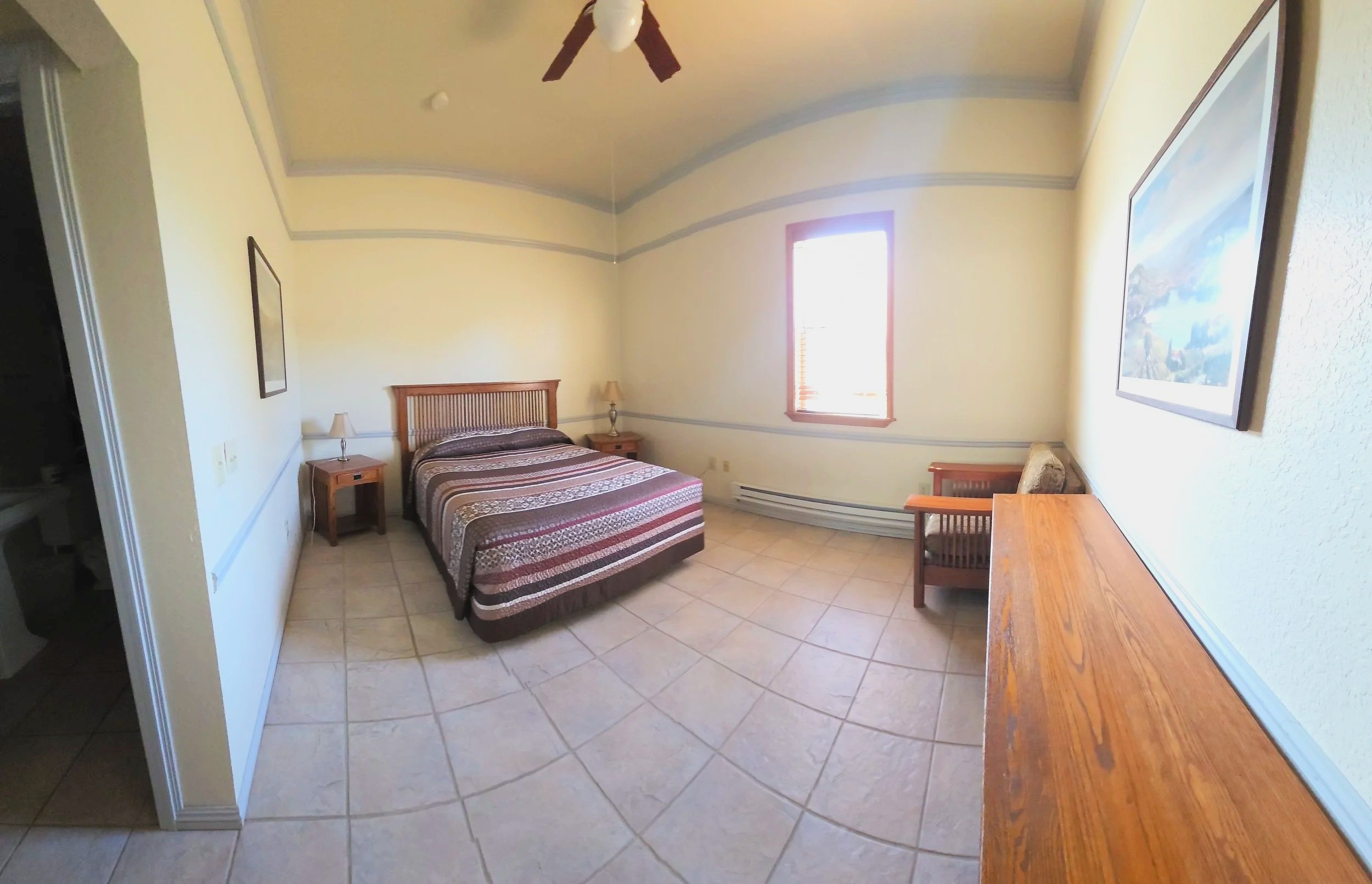 A bedroom with a bed, two nightstands with lamps, a wooden bench, a window with blinds, a piece of furniture with a wooden surface, and a framed picture on the wall.