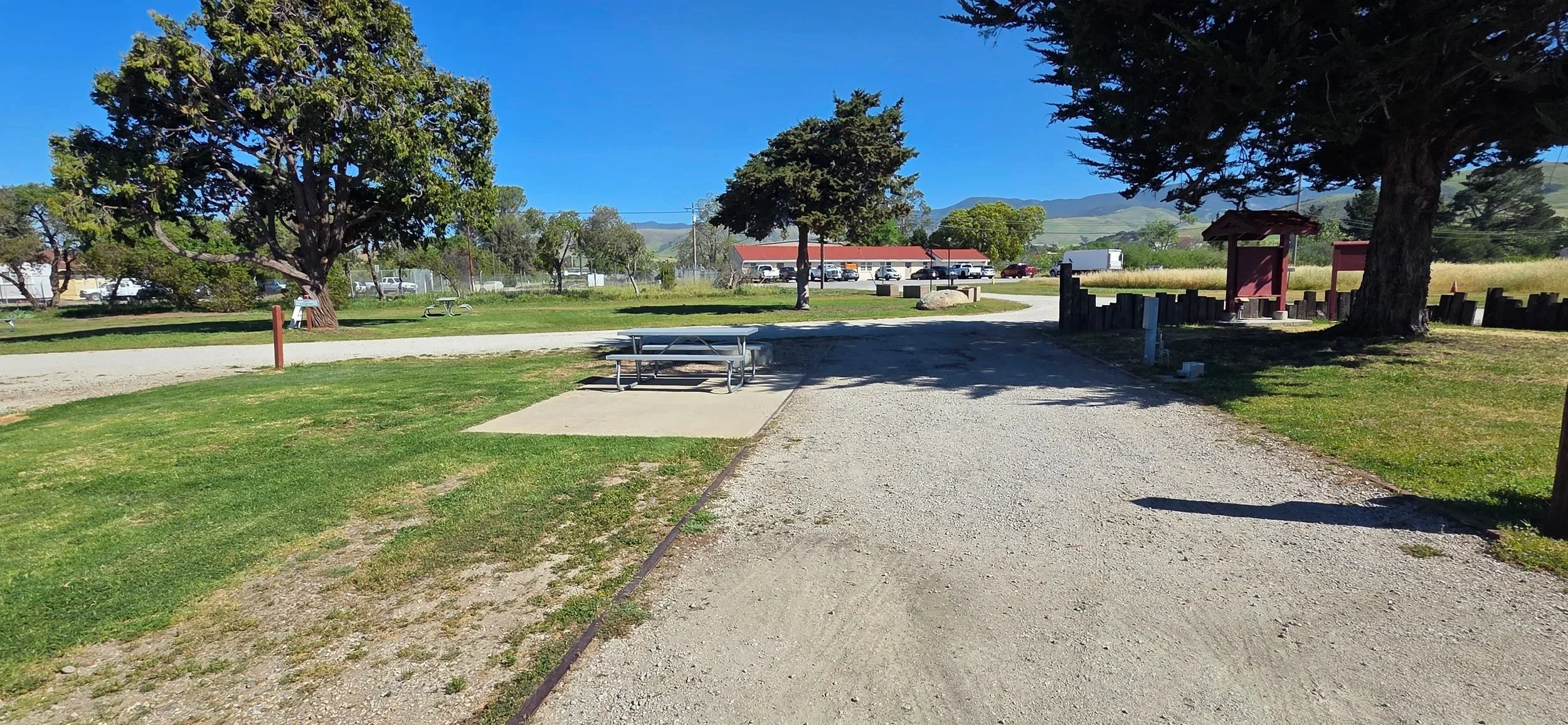 A park with grass, trees, a picnic table, a gravel path, and a viewing area with a small shelter.