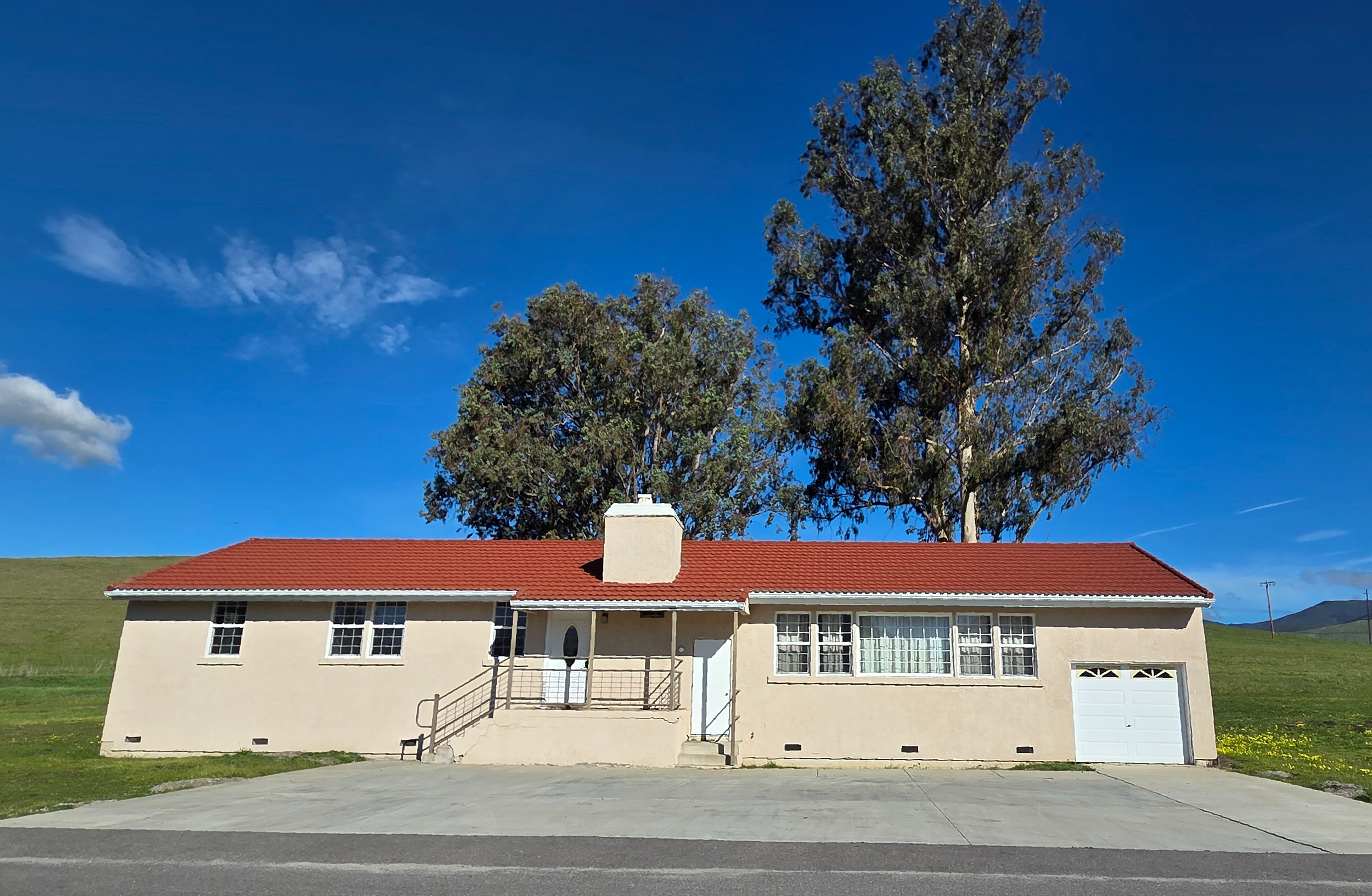 Single-story house with light-colored exterior, red tiled roof, front porch with railing, garage on the right, concrete driveway, green grassy area, large trees behind the house, blue sky with scattered clouds.