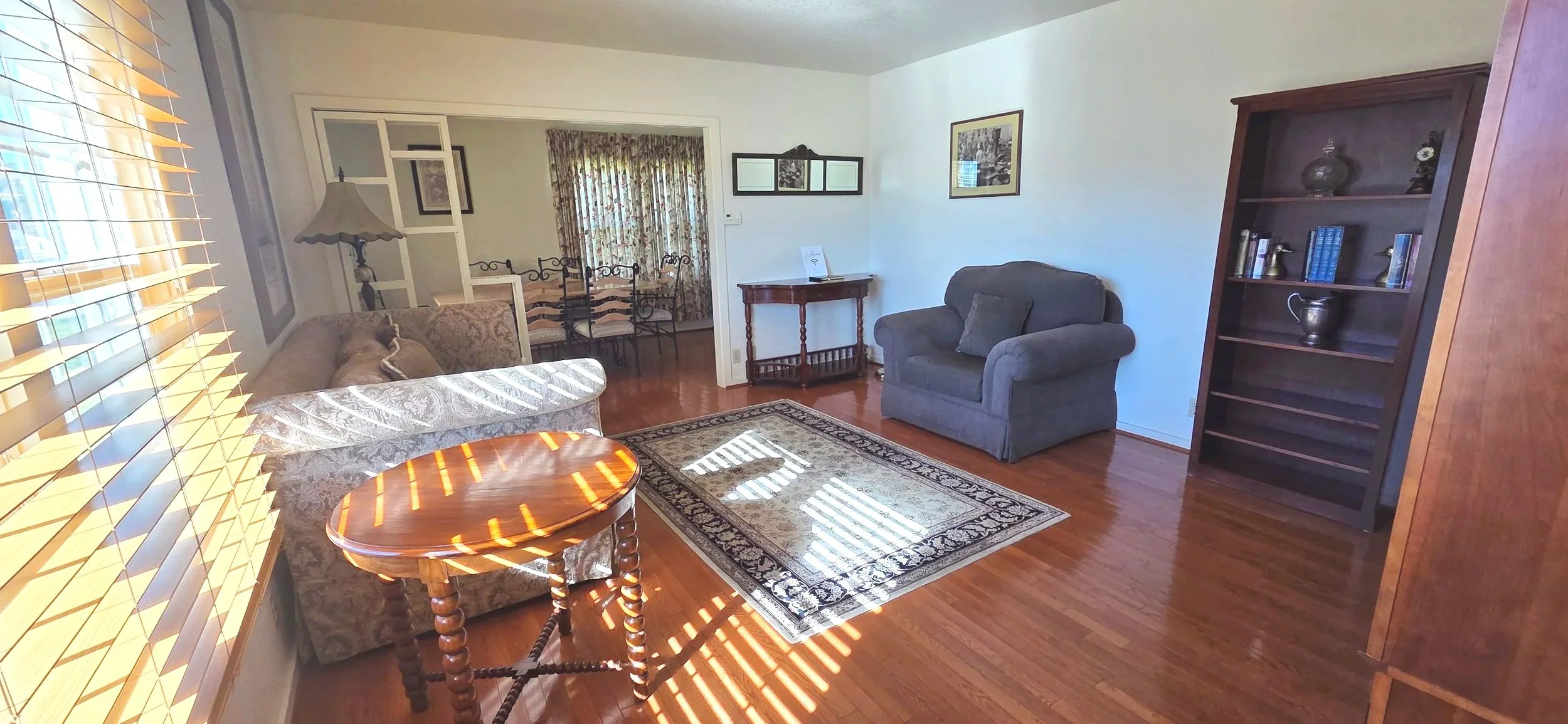 A cozy living room with hardwood flooring, a floral-patterned sofa, and a dark armchair. Sunlight filters through window blinds, casting striped shadows. A patterned area rug is in the center, and a wooden side table is near the sofa. In the background, a dining area with a table and chairs is visible, separated by a partial wall with a decorative mirror on the wall.