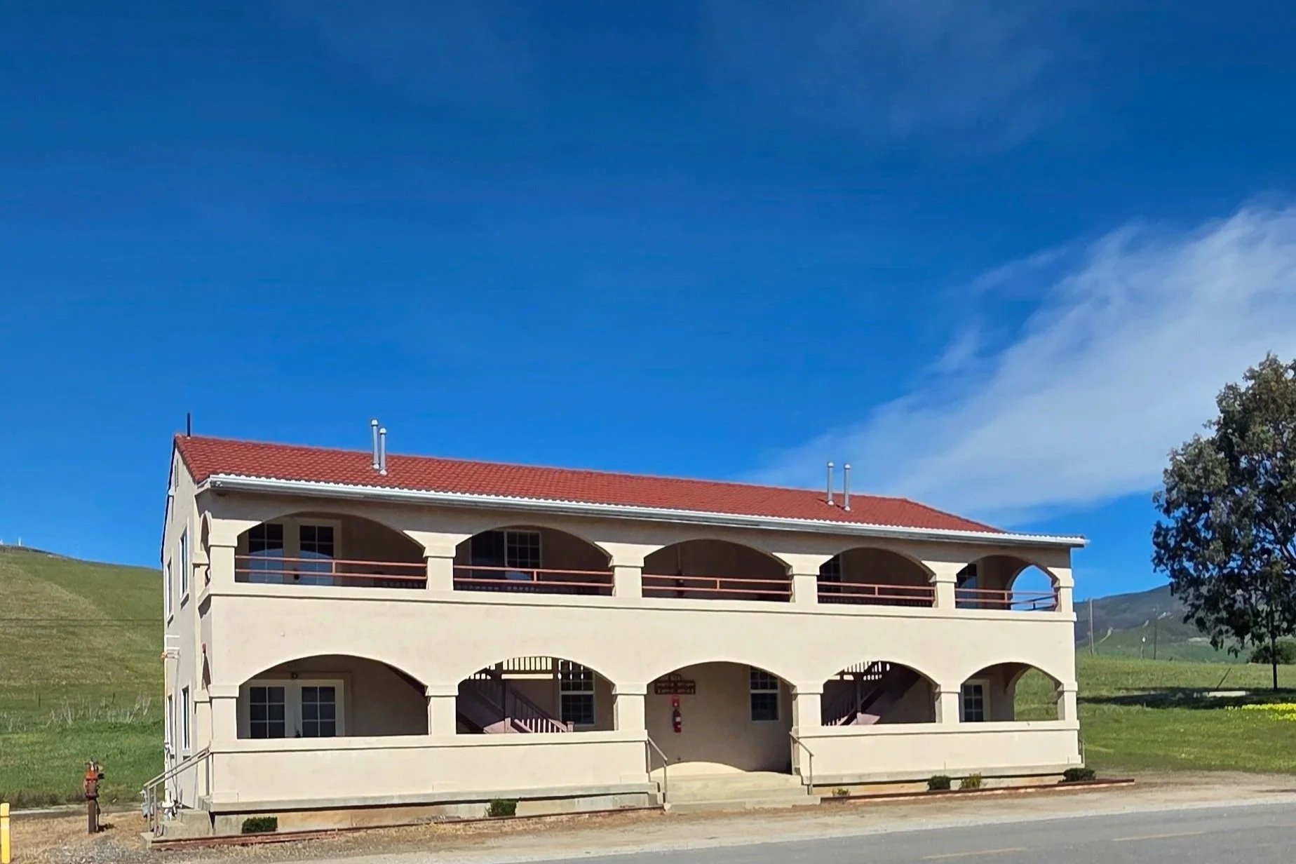 A two-story building with a red-tiled roof and cream-colored walls, featuring arched balconies on both floors and a staircase entrance in the center, set against a backdrop of green rolling hills and a bright blue sky.