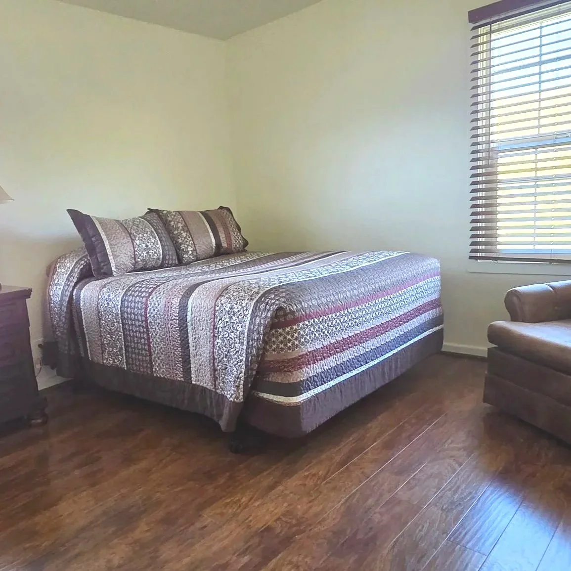 A bedroom with a large bed covered in a patterned quilt and pillows, a window with blinds on the right, and a leather armchair on the lower right corner.