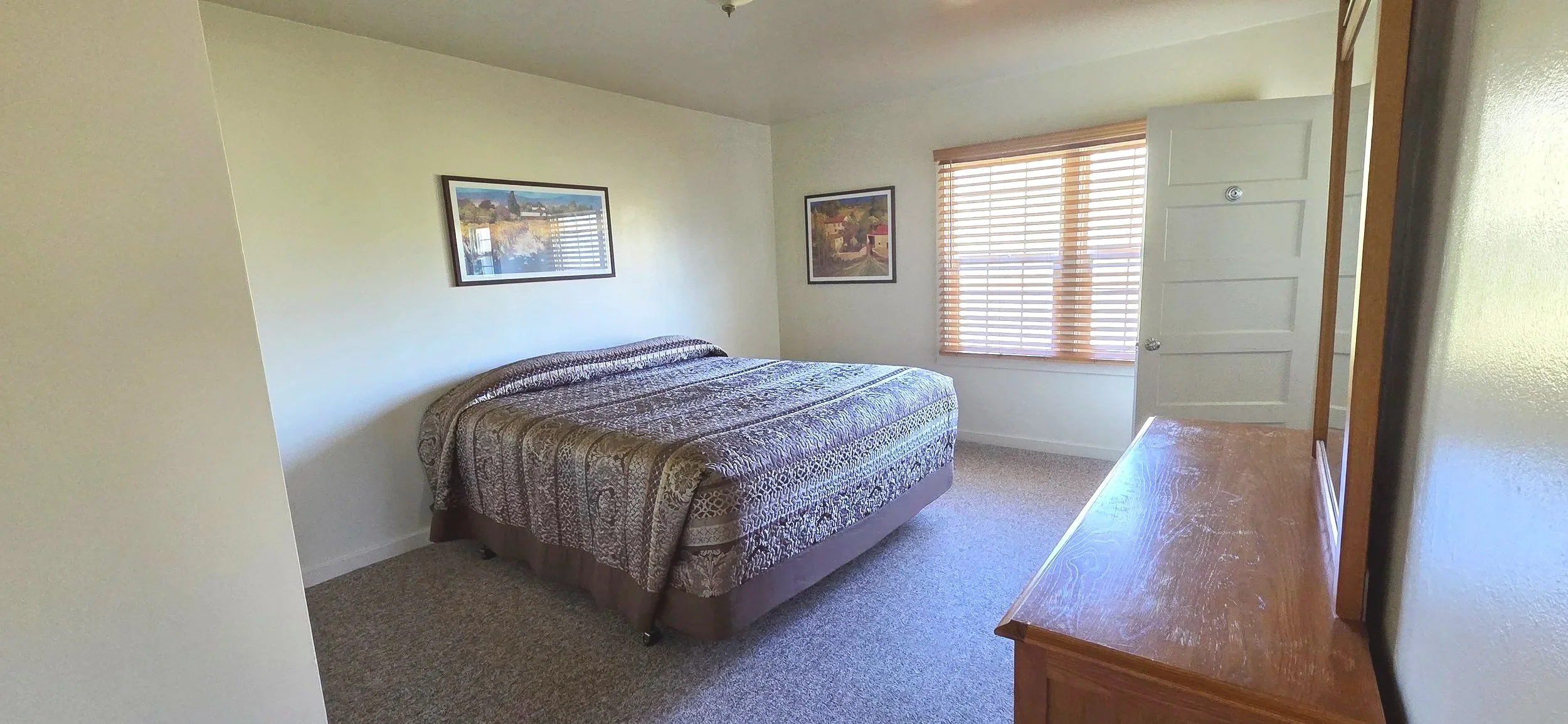 A bedroom with a bed against the wall, two framed pictures above, a window with blinds, and a wooden dresser on the right.