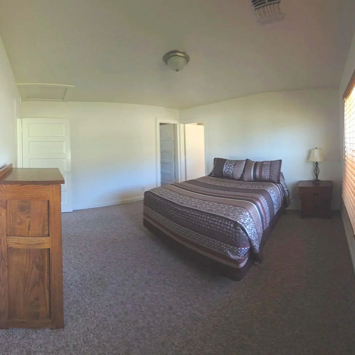 A bedroom with a bed featuring a multicolored patterned bedspread and pillows, a wooden nightstand with a lamp, a wooden dresser, white walls, a window with blinds, and carpeted flooring.