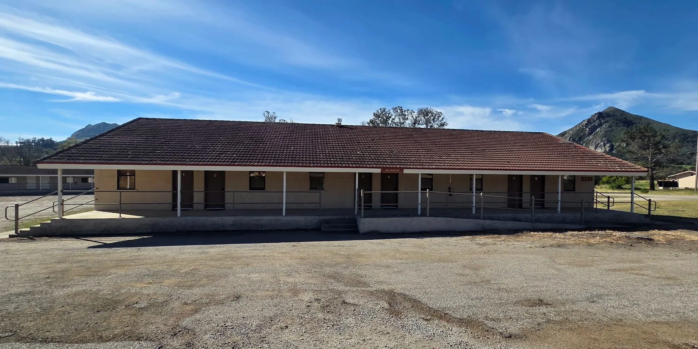 Single-story beige building with a red tile roof, multiple doors and small windows, accessible by stairs and designed for handicap access, set against mountains and a clear blue sky.