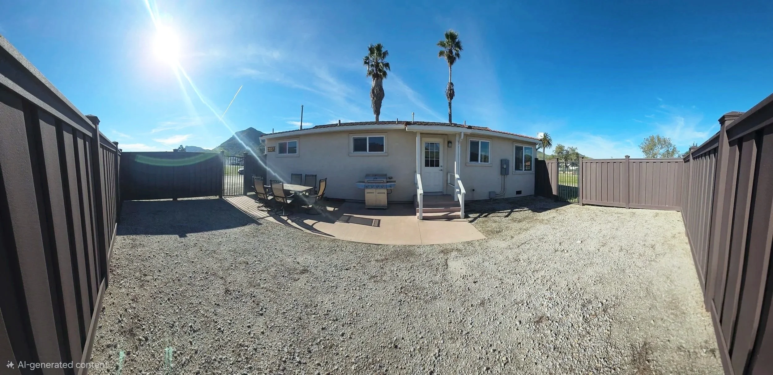 Fenced backyard with gravel ground, a small patio area with a dining table and chairs, a propane grill, a white house with steps leading up to a door, several tall palm trees, and a mountain in the background under a clear blue sky with the sun shining brightly.