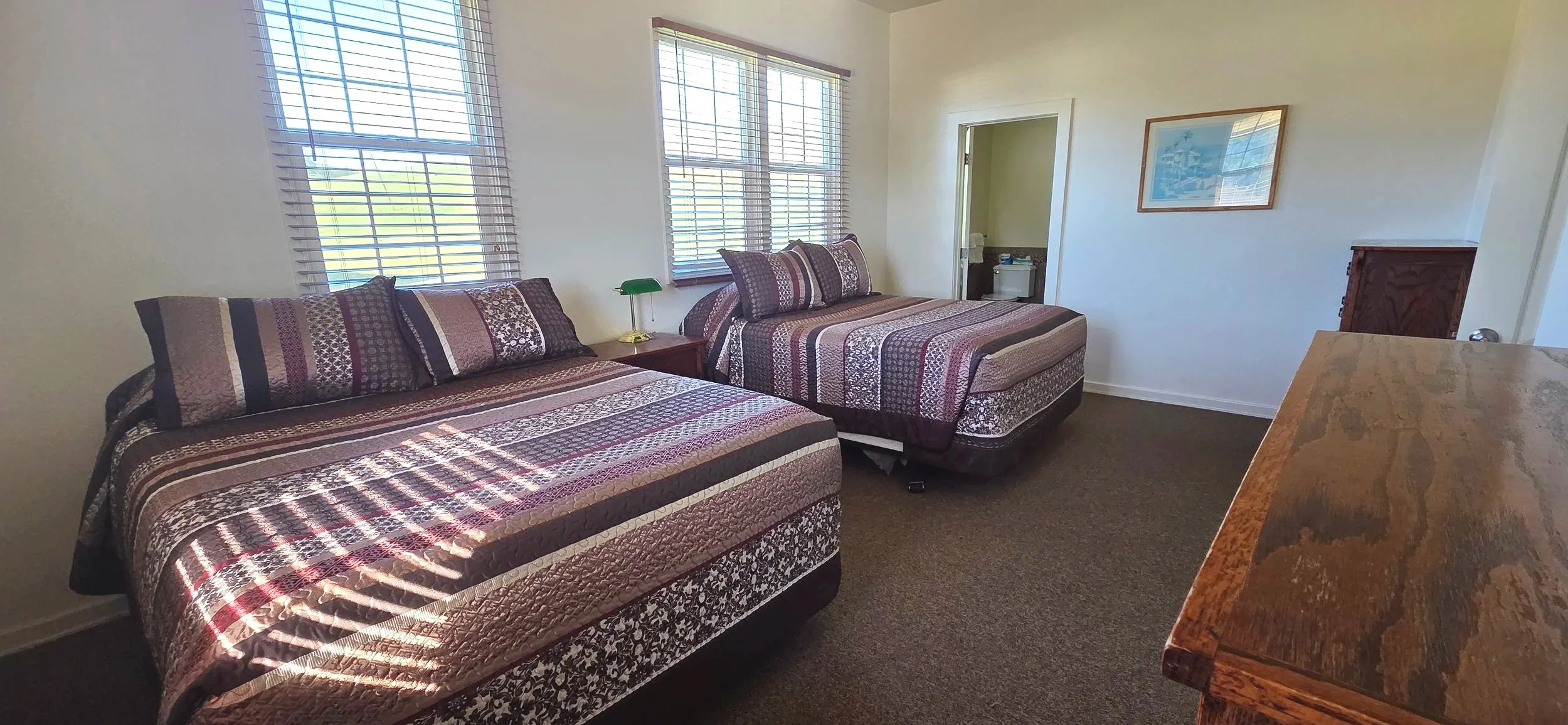 A bedroom with two double beds with striped quilt covers, placed side by side. Each bed has three pillows with patterned pillowcases. There are three windows with blinds on the left wall, allowing sunlight into the room. A small wooden nightstand with a green desk lamp sits between the beds. To the right, there is a doorway leading to a bathroom and a wooden dresser. A framed picture hangs on the wall above the dresser. A wooden dresser with a rich grain pattern is partially visible on the right side of the image.