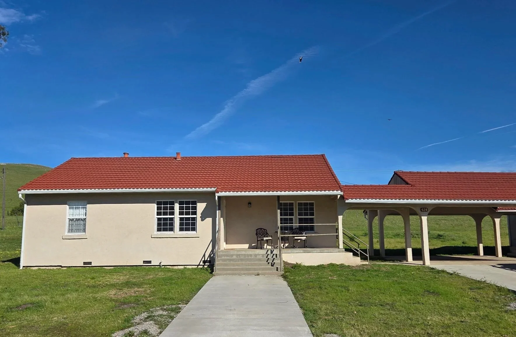 Single-story house with a red tiled roof, beige exterior walls, and a small front porch with seating, set against a bright blue sky with a hillside in the background.