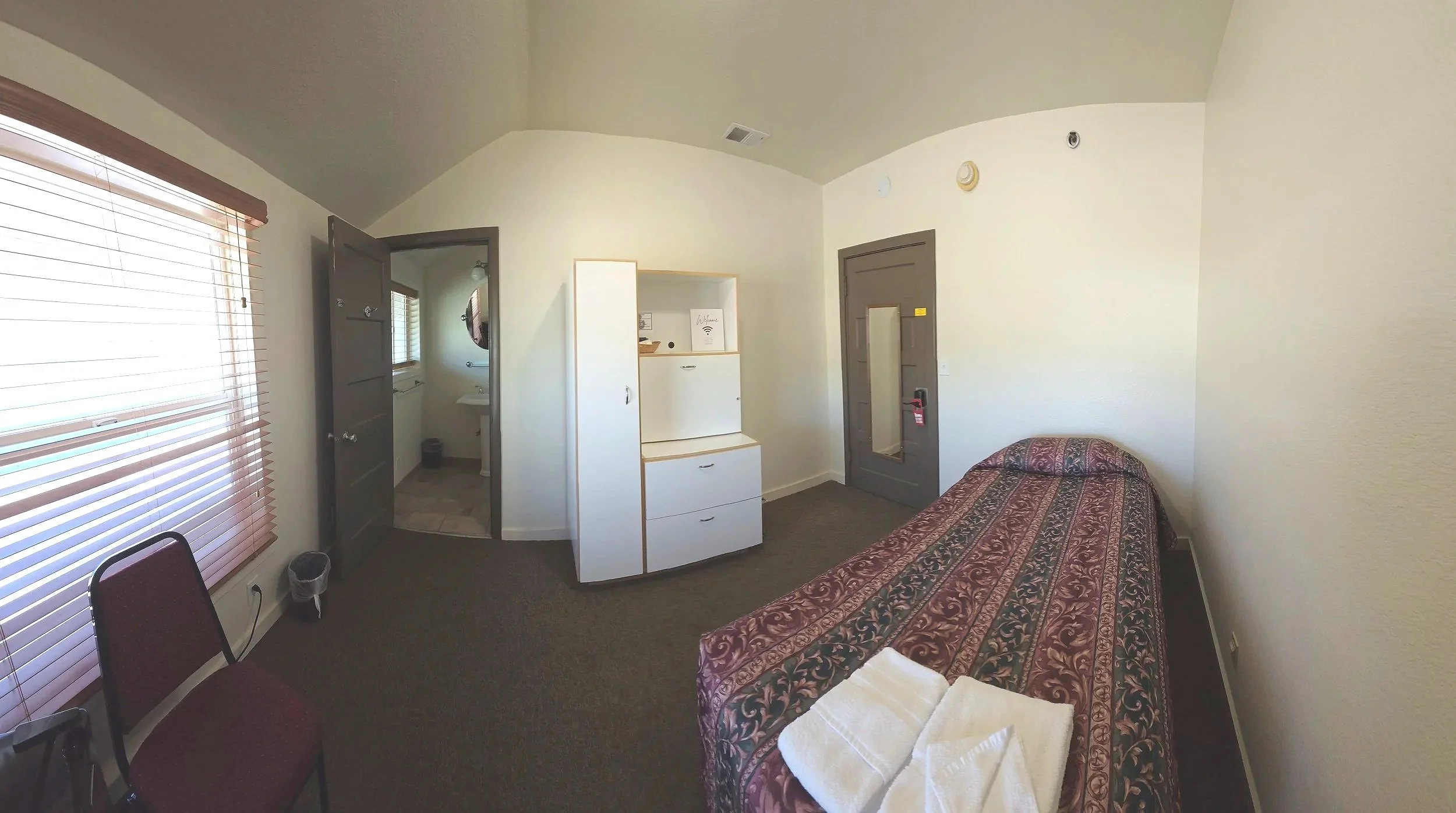 Room with a single bed covered with a patterned blanket, a chair near a window with blinds, a white storage cabinet, and a door leading to a bathroom visible through an open door.