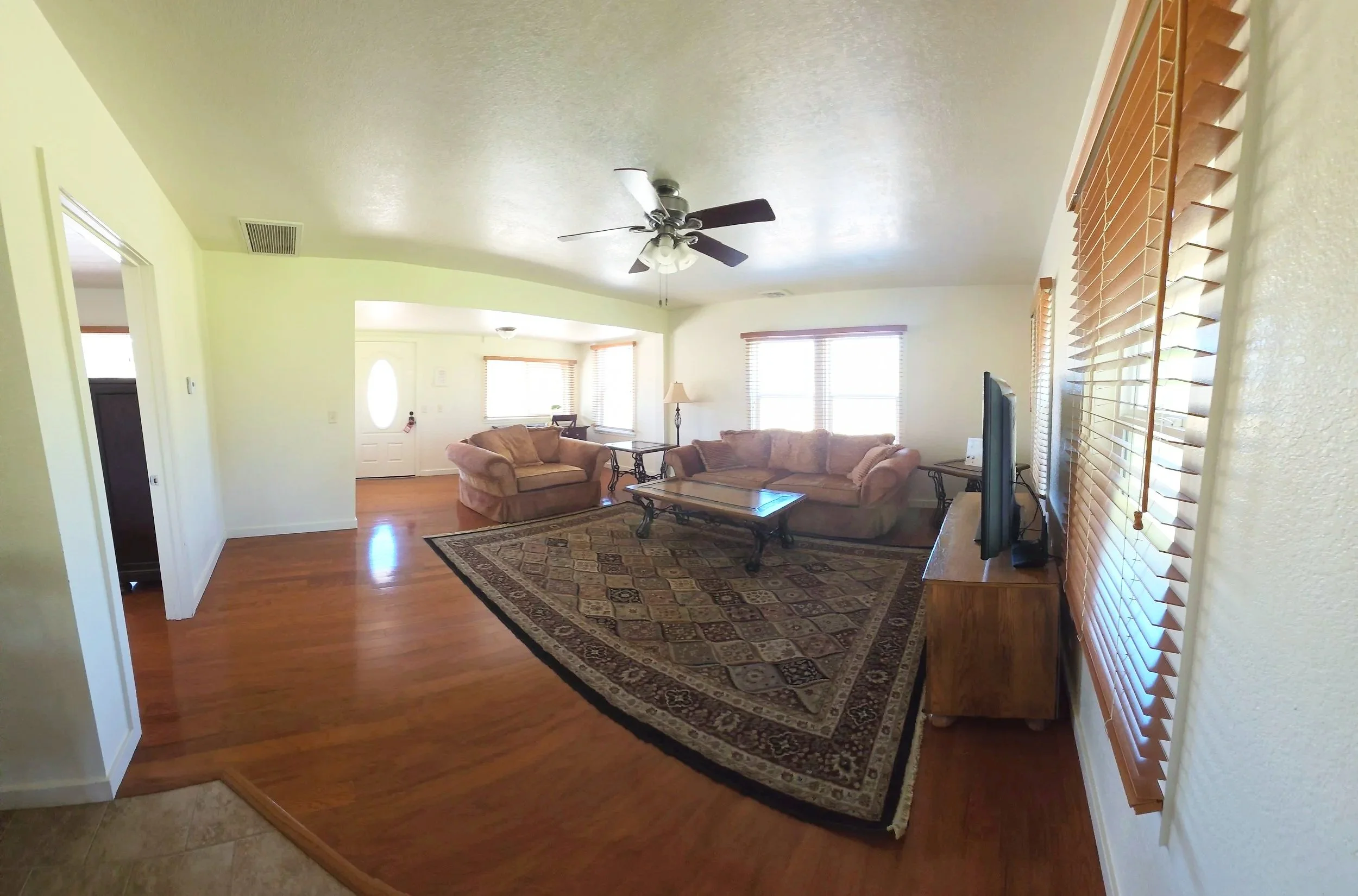 Living room with two sofas, a coffee table, area rug, television on a wood stand, ceiling fan, and multiple windows with wooden blinds.