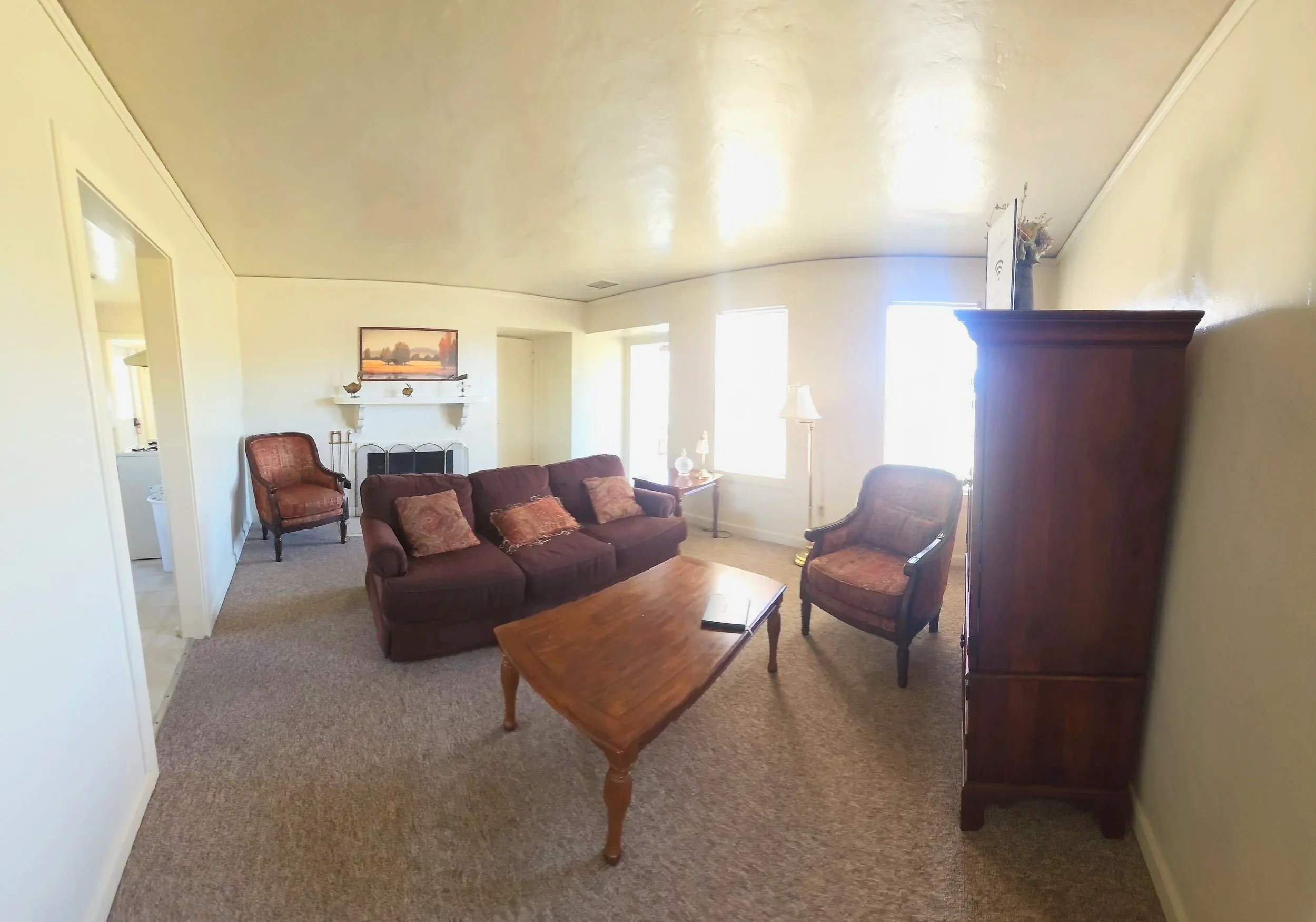 Living room with a maroon sofa and matching armchair, wooden coffee table with remotes, a wooden cabinet, a fireplace with decorative items, and large windows letting in natural light.