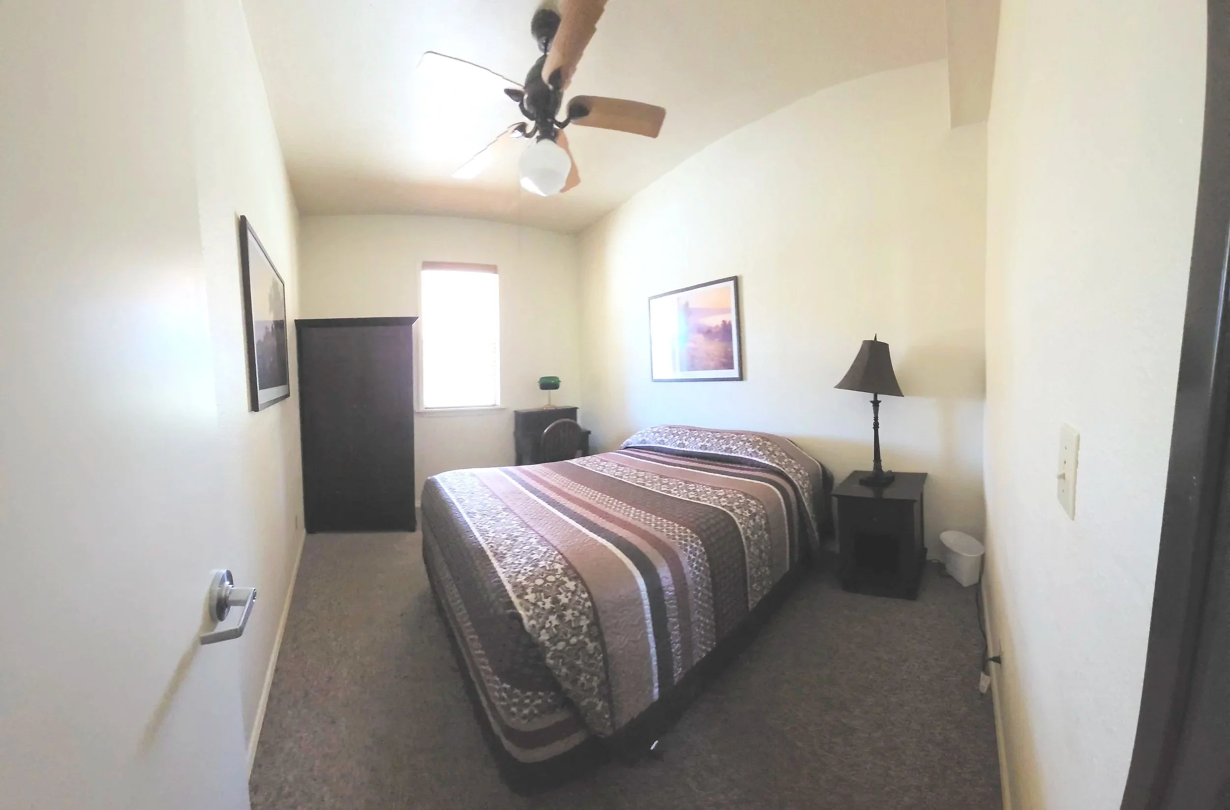 A cozy bedroom with a bed, nightstand, table lamp, framed artwork, window, ceiling fan, dark dresser, and carpeted floor.