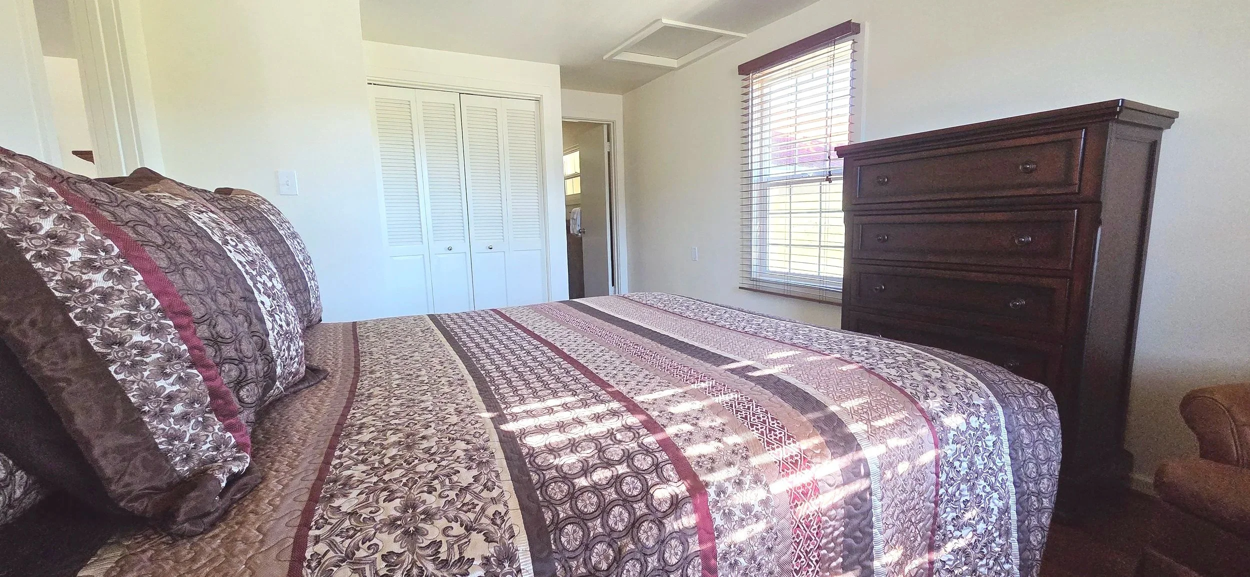 A bedroom with a bed featuring a patterned bedspread and pillows, a window with blinds, a brown dresser, and a closet with white double doors.