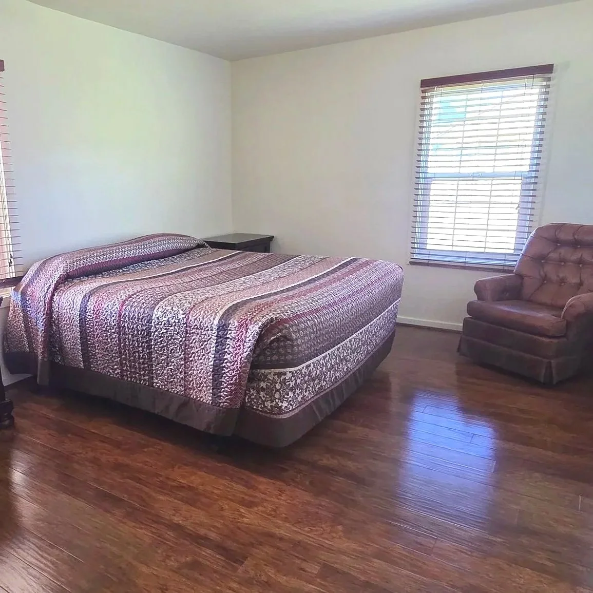 A bedroom with a bed covered in a patterned quilt, a small bedside table, a window with blinds, and an upholstered armchair in the corner.