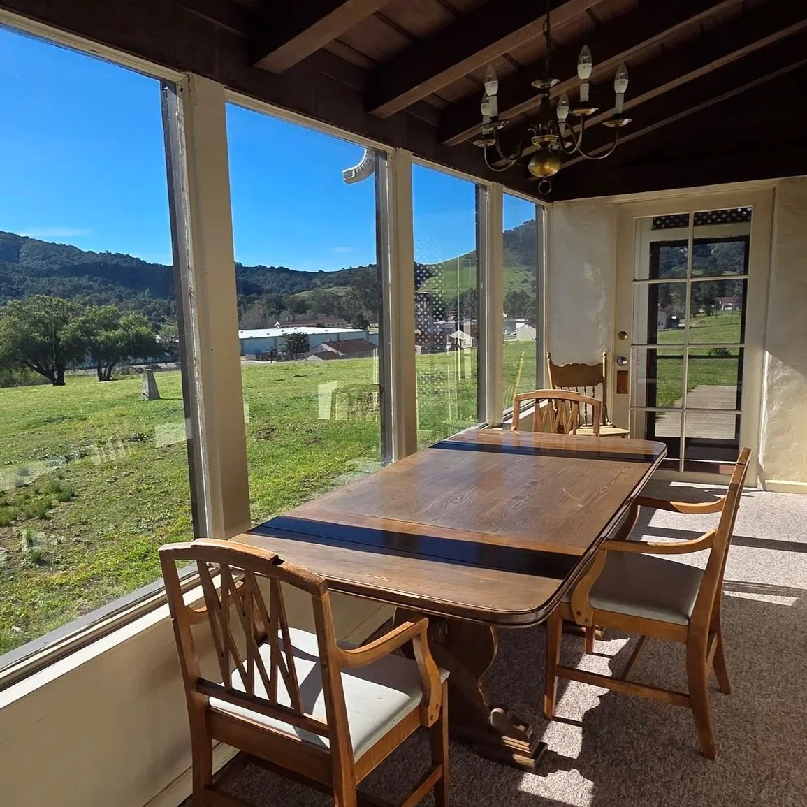 Sunlit dining area with wooden table and chairs, large windows showing a green field, trees, and distant mountains, a chandelier hangs from a wooden ceiling, and a glass door leads outside.
