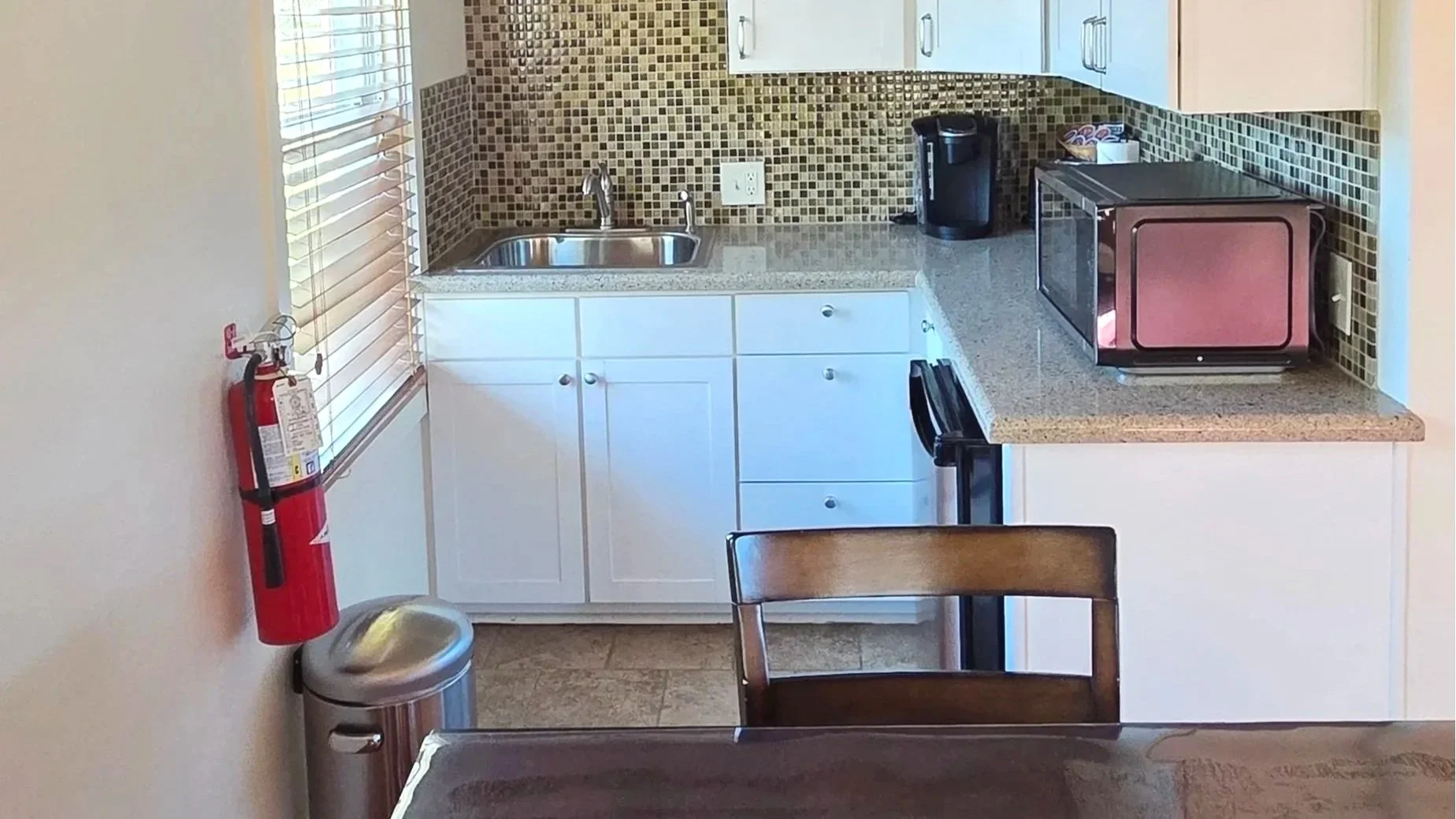 Small kitchen with white lower cabinets, a granite countertop, and a mosaic tile backsplash. There is a stainless steel sink, a coffee maker, a microwave, and a window with blinds.
