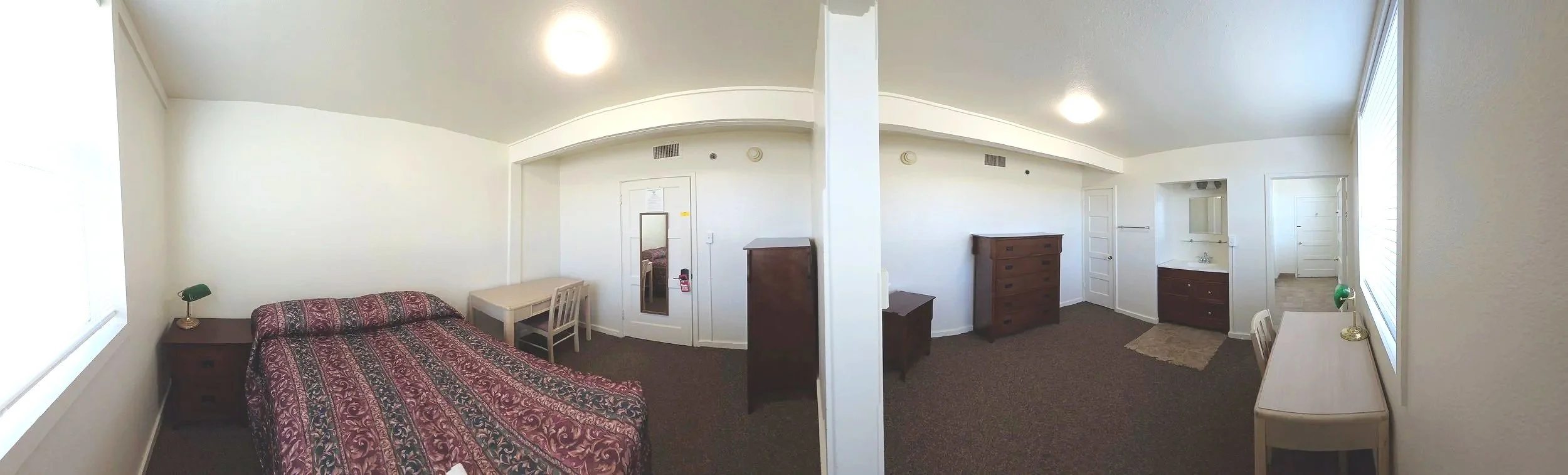 Interior of a simple, furnished room with a bed, nightstand, desk, mirror, dresser, and a small bathroom area with a sink, all in a neutral color scheme.