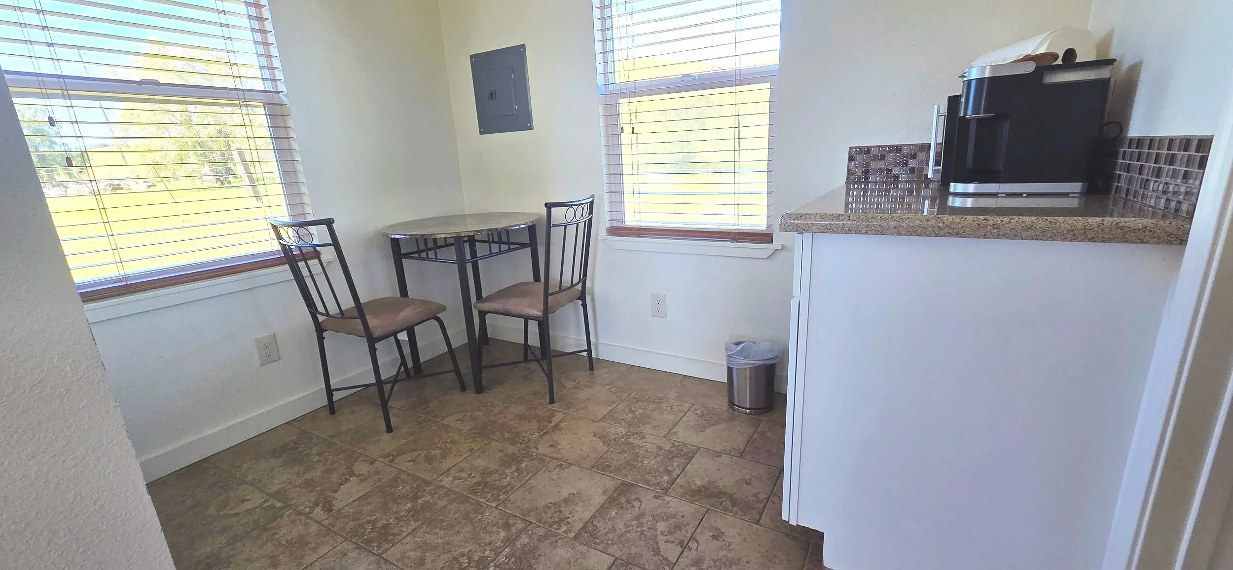 Small dining area with two windows showing green fields outside. There is a small round table with two chairs. The chairs have metal frames and cushioned seats. A countertop with a coffee maker and a small backsplash is visible. A trash can is placed next to the counter. The floor has brown tiles, and the walls are painted light.