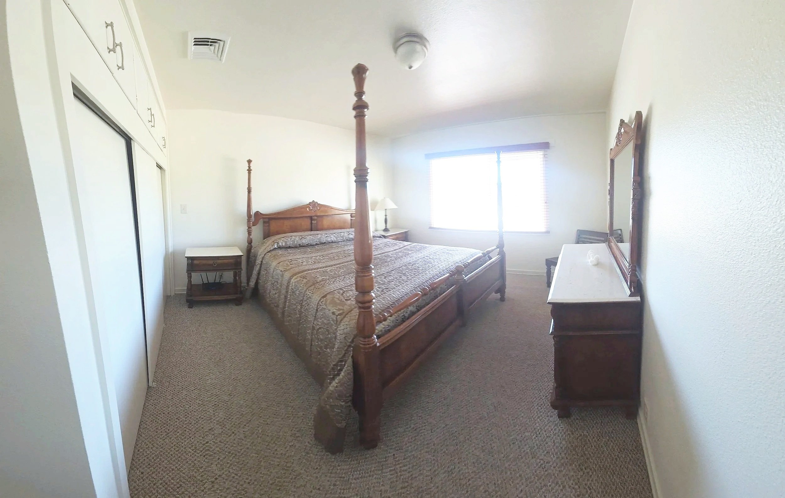 A bedroom with a wooden four-poster bed, window with blinds, nightstand with lamp, dresser with mirror, and closet with sliding doors.