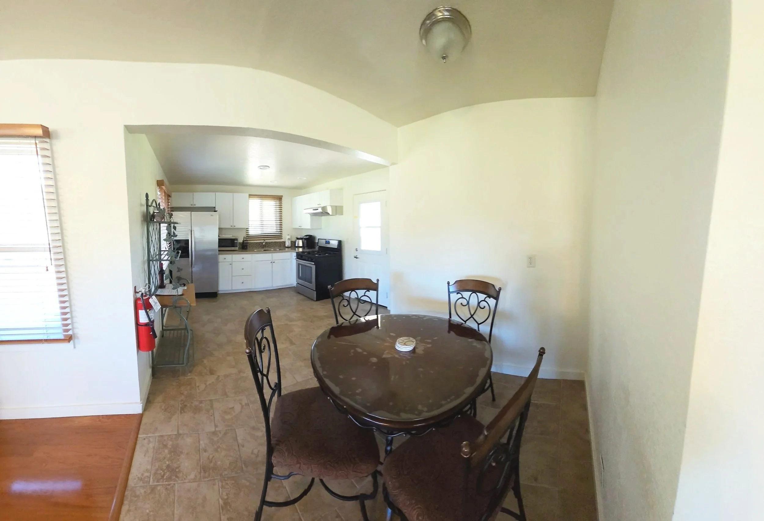 Dining area with a round table and four chairs, adjacent to a kitchen with white cabinets, stainless steel refrigerator, stove, microwave, and windowed walls, in a home interior.
