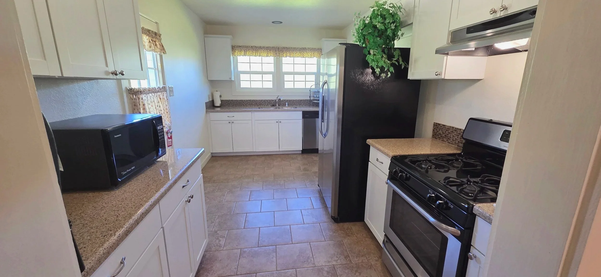 A kitchen with white cabinets, beige countertops, a microwave, a stove, and a refrigerator, with a window above the sink and a potted plant on top of the refrigerator.
