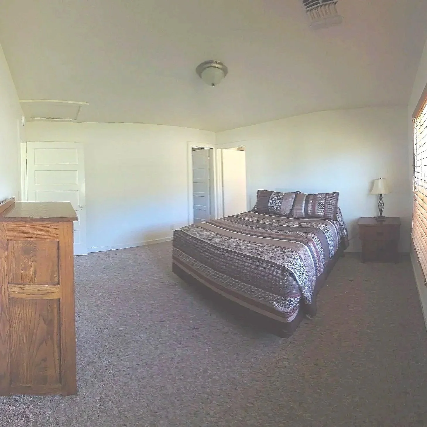 A bedroom with a bed covered in a striped quilt and matching pillows, a wooden nightstand with a lamp, a wooden dresser, and a window with closed blinds. White walls and carpeted floor.