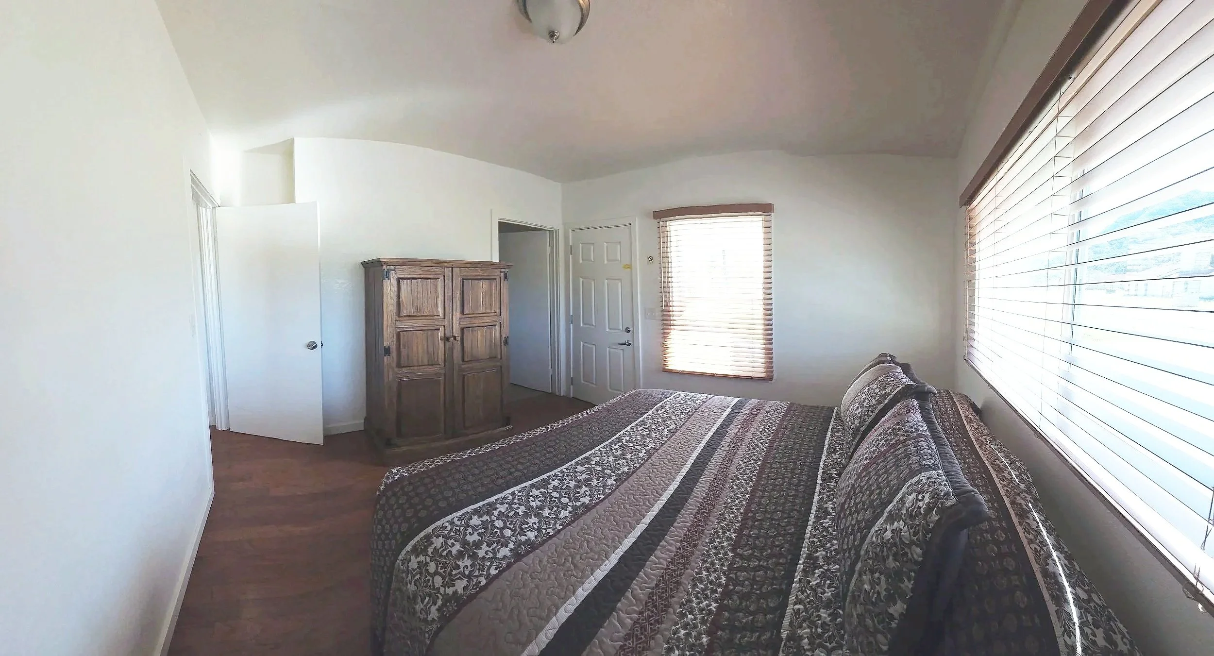 A bedroom with a large bed covered in a patterned quilt, two pillows, and wooden horizontal blinds on two windows letting in natural light. There's a wooden armoire near the door and hardwood flooring.