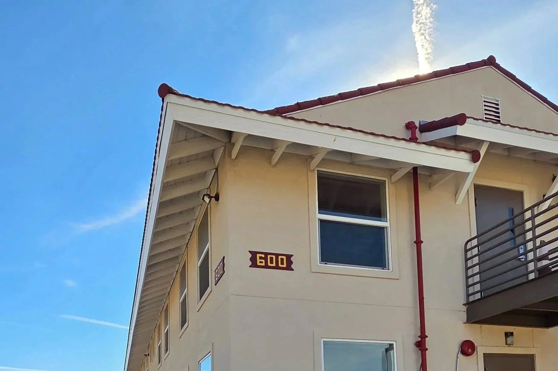 Close-up view of the upper corner of a light yellow residential building with a red tile roof, windows, a balcony, and a fire alarm. The blue sky with some clouds is visible in the background.
