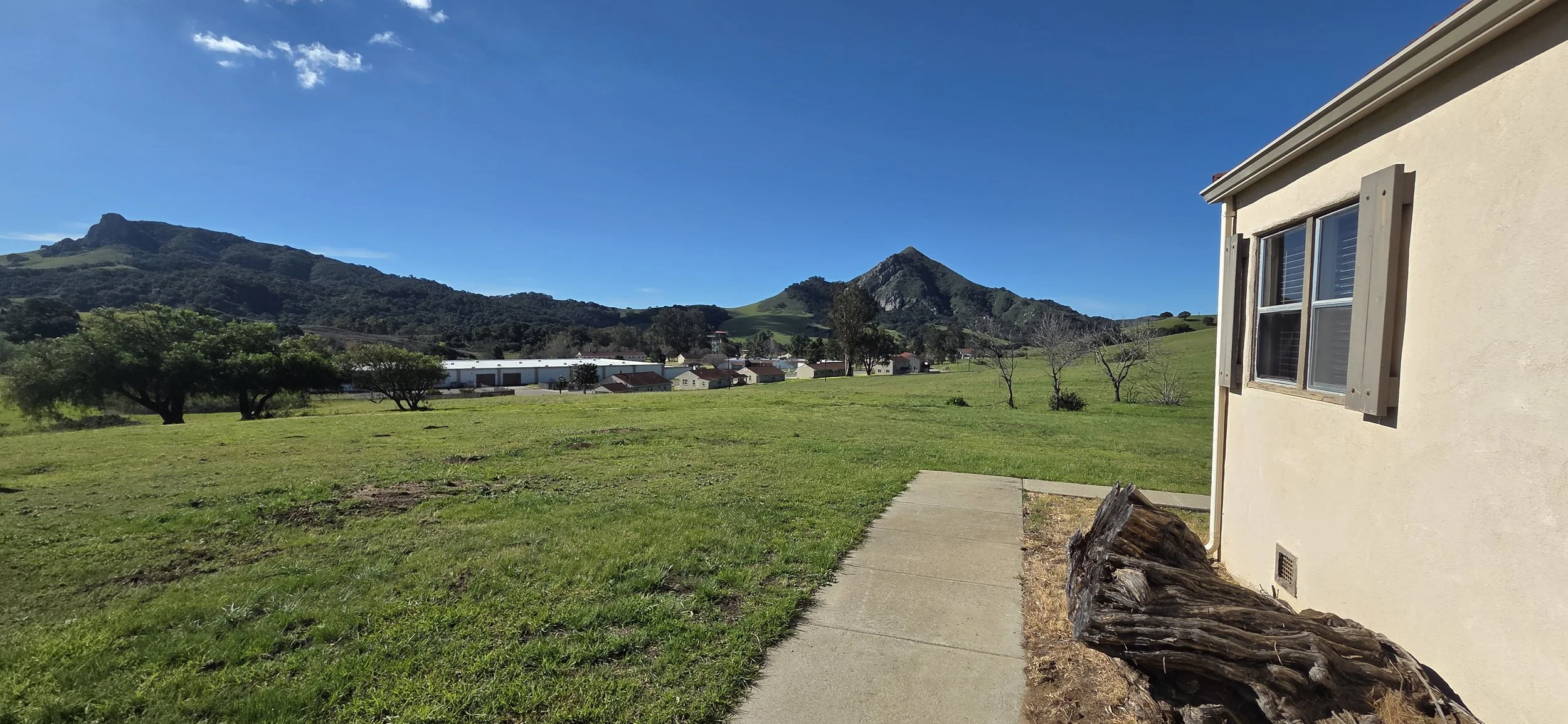 A side view of a house with a window and wooden shutters, a concrete walkway, a large piece of driftwood, and a grassy yard with a few trees, set against distant green mountains under a bright blue sky.