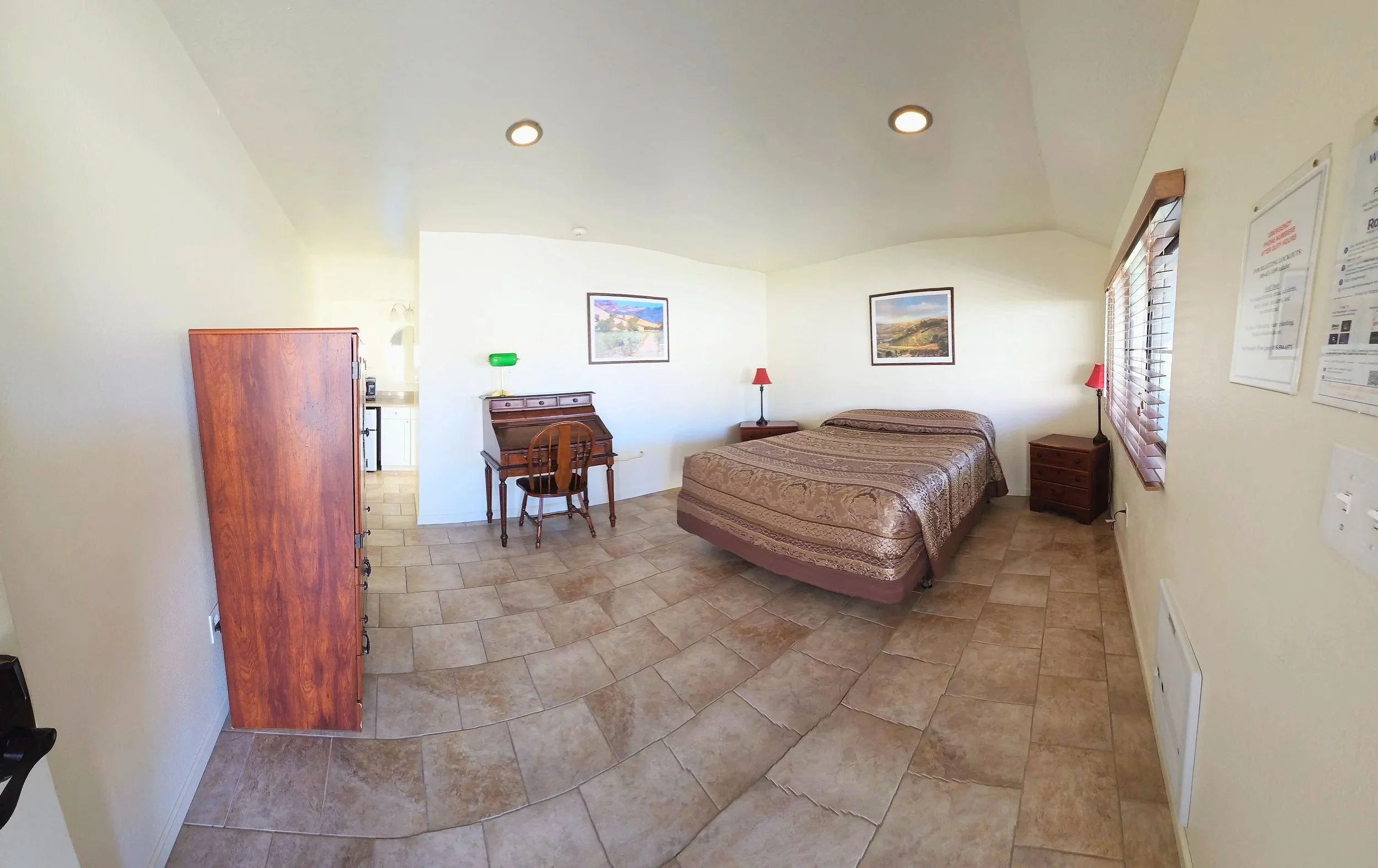 Hotel room with a bed, side tables, lamps, a wooden desk, a chair, and a window with blinds.