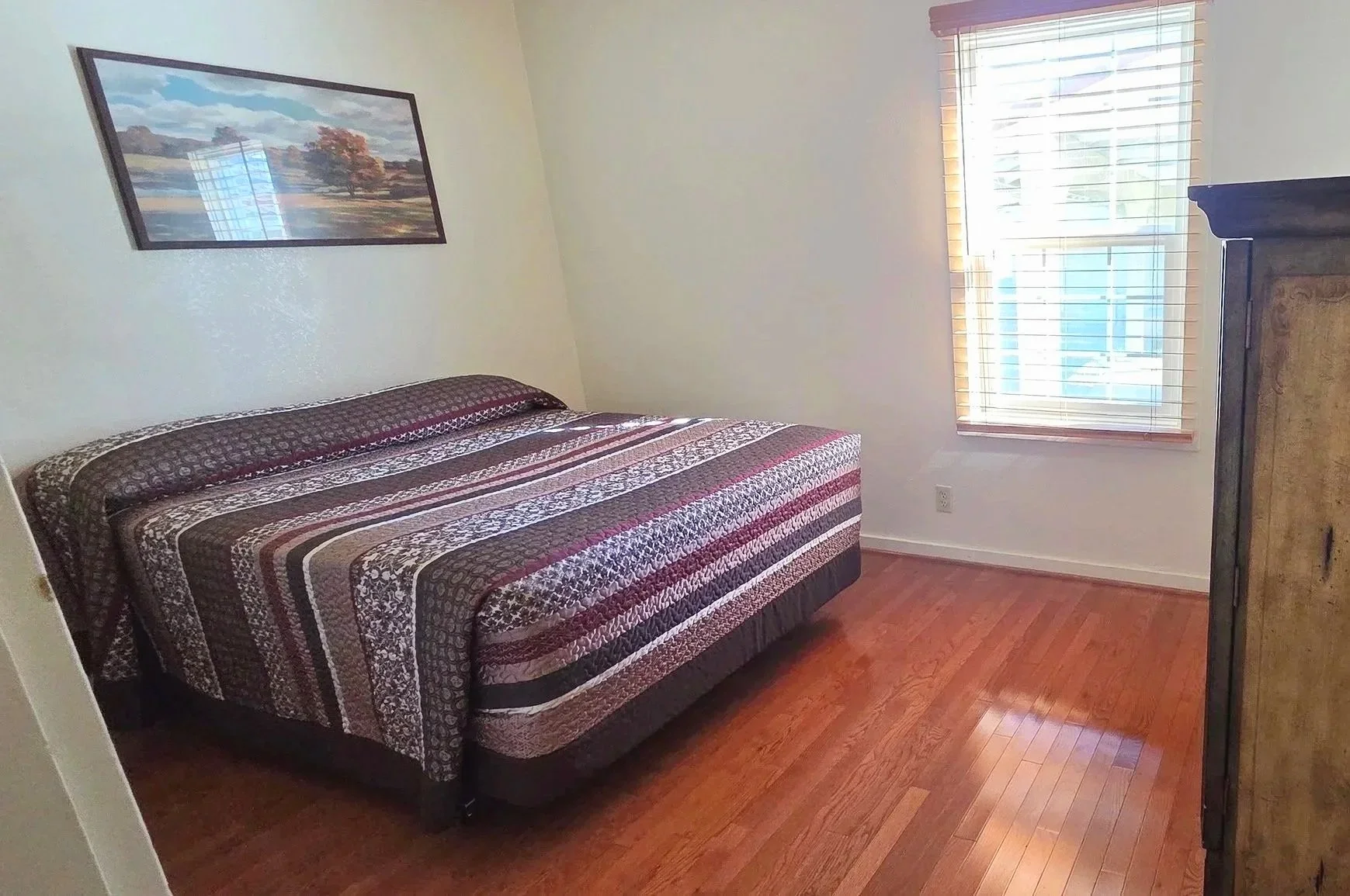 Bedroom with a bed having a striped blanket, a window with wooden blinds, a framed landscape painting above the bed, and a wooden dresser.