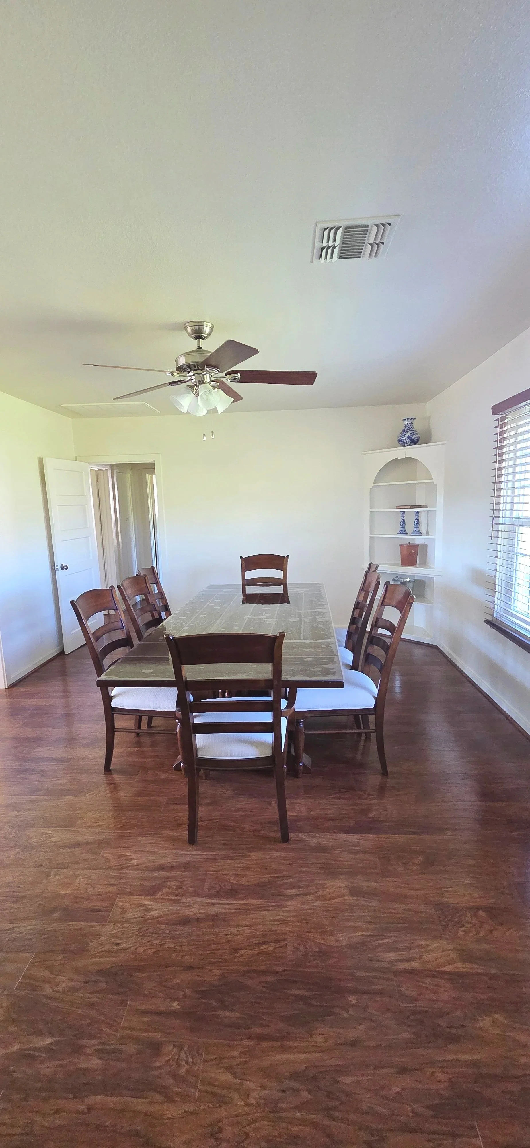 Dining room with a wooden table and six chairs, a ceiling fan with lights, a window with blinds, and a white built-in shelf with decorative ceramics, on a hardwood floor.