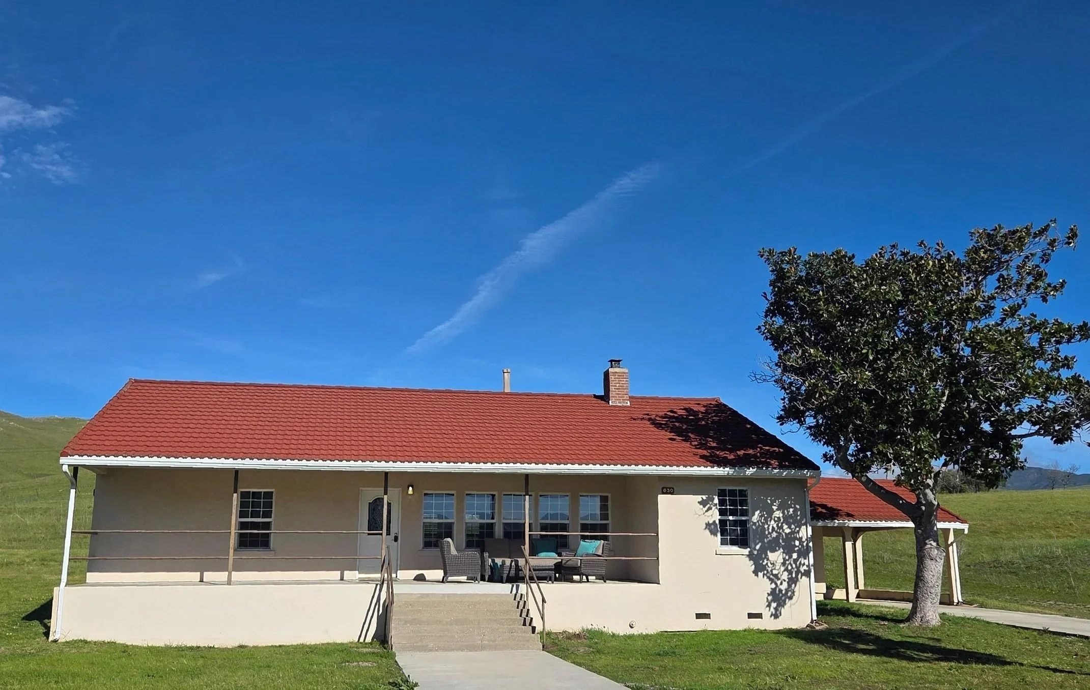 A house with a red tiled roof, a porch with outdoor seating, trees, and a grassy landscape under a blue sky.