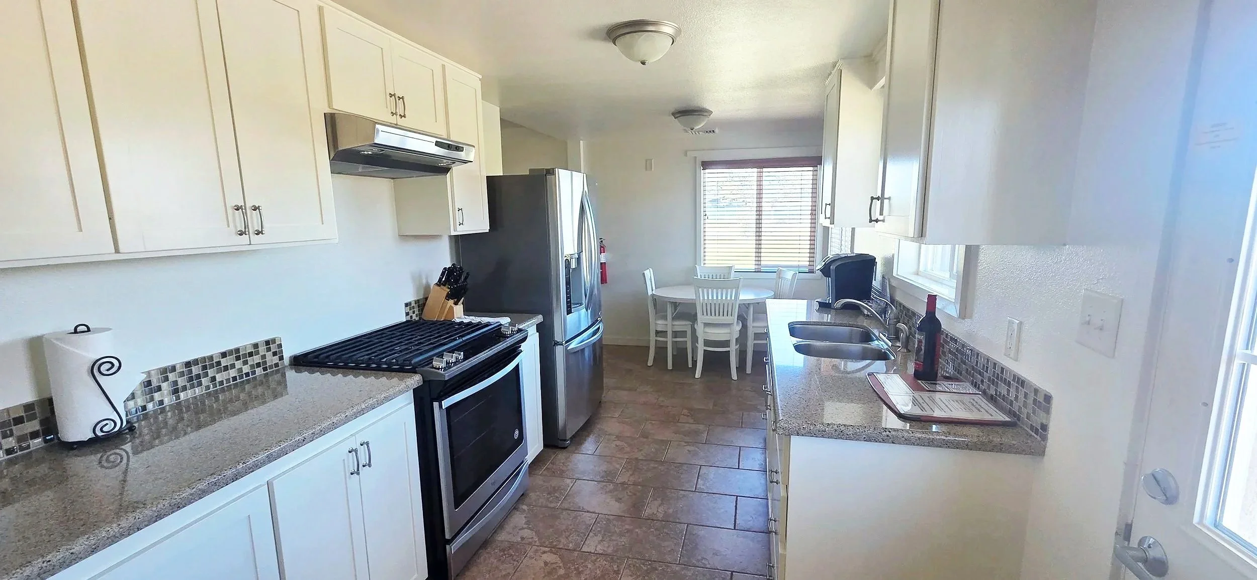 Kitchen with white cabinets, a granite countertop, stainless steel appliances including a refrigerator and stove, a round dining table with four chairs, and a window with blinds overlooking a grassy area.