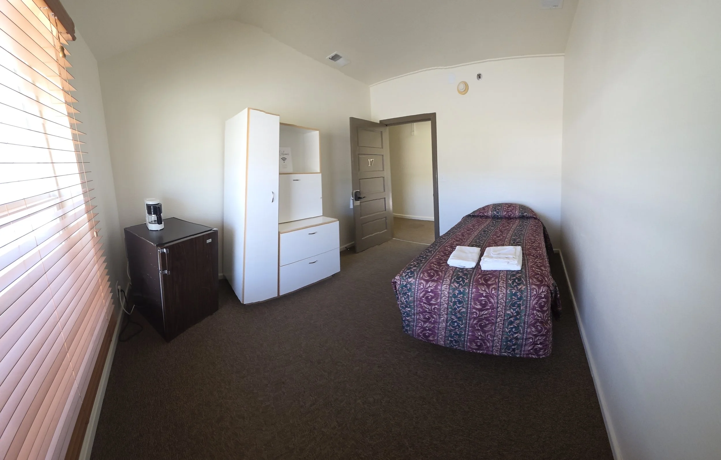 Hotel room with single bed, folded white towels, window with blinds, small refrigerator, white cabinet, and open door.