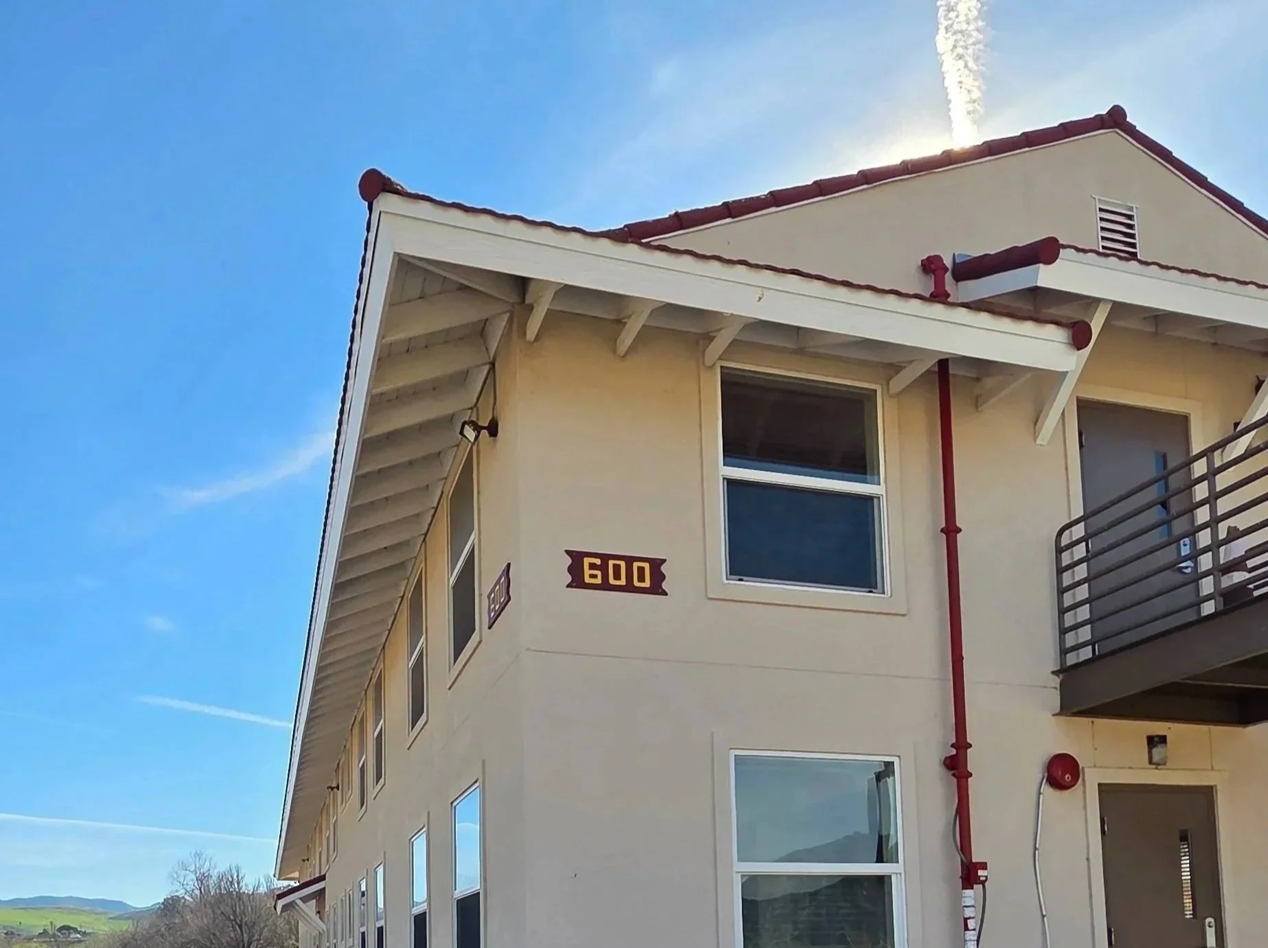 Close-up of a corner of a beige building with white trim, multiple windows, a red pipe running vertically, and a sign with the number 600. The sky above is clear and bright.
