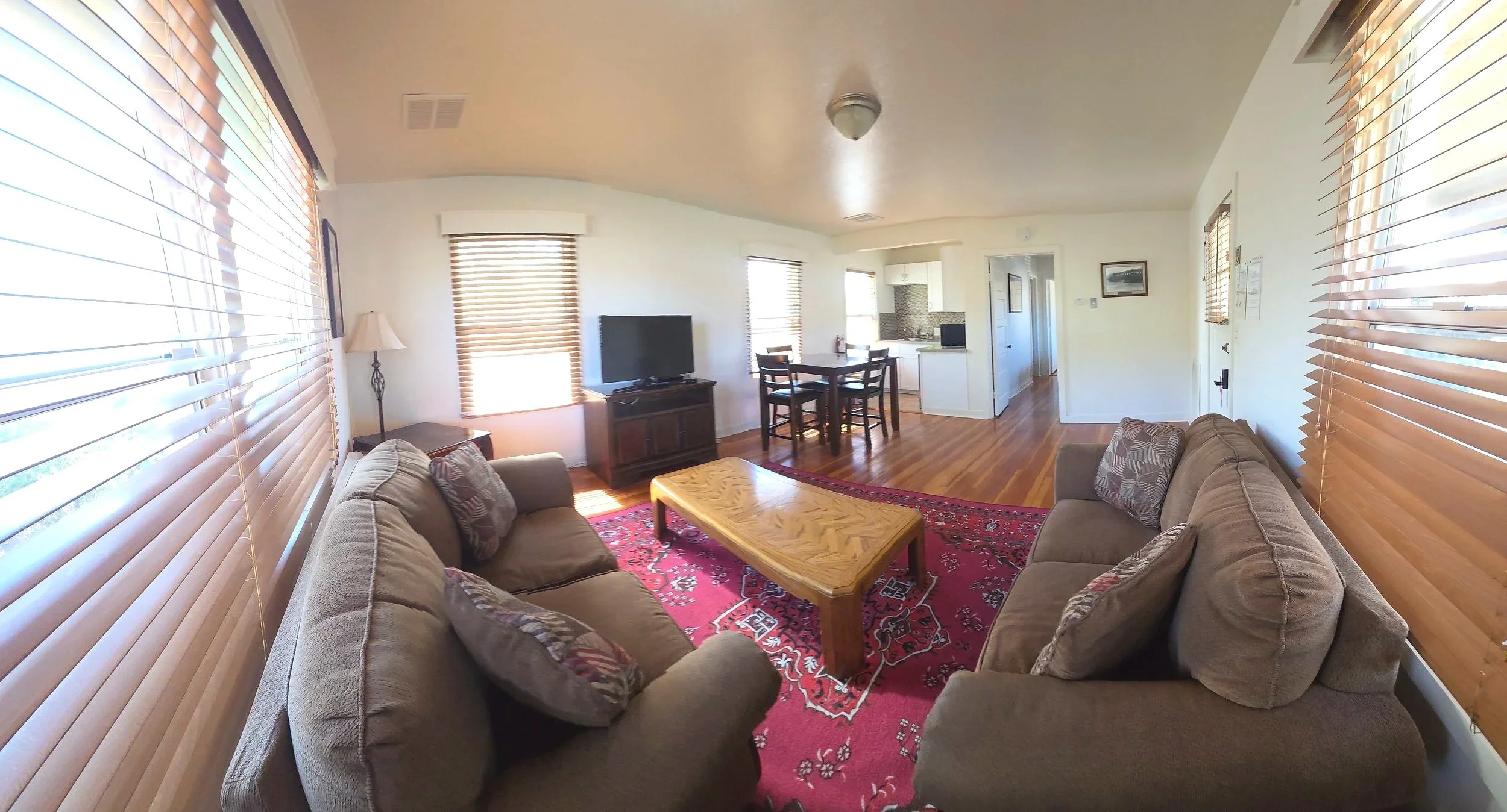 Living room with hardwood floors, beige sofas, a red patterned rug, and wooden blinds on the windows.