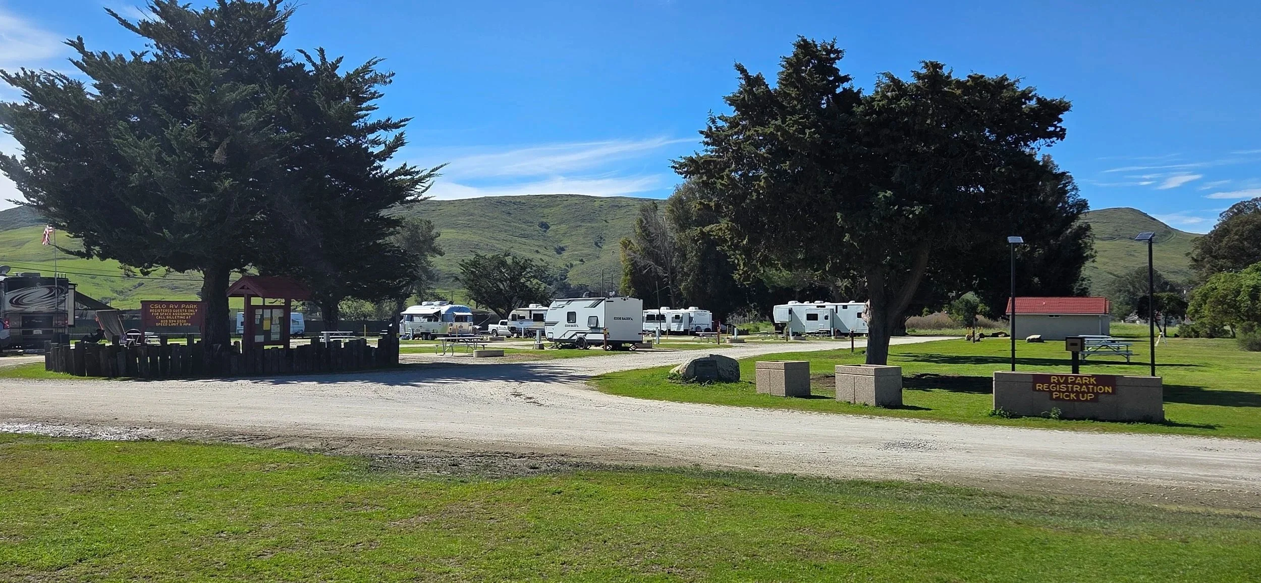 A campground with trees, RVs, and a gravel road under a clear blue sky. Signage indicates RV park registration and pickup area.