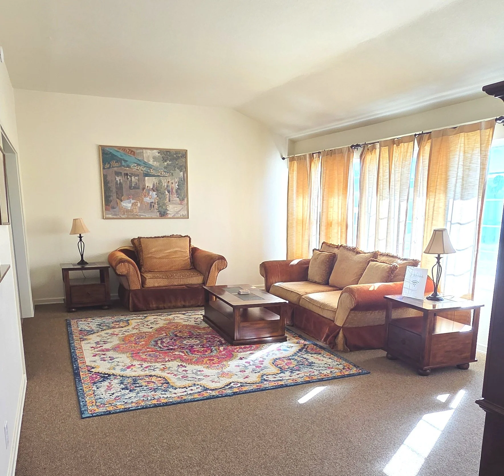 Living room with two sofas, a coffee table, side tables with lamps, a colorful area rug, and large windows with beige curtains.