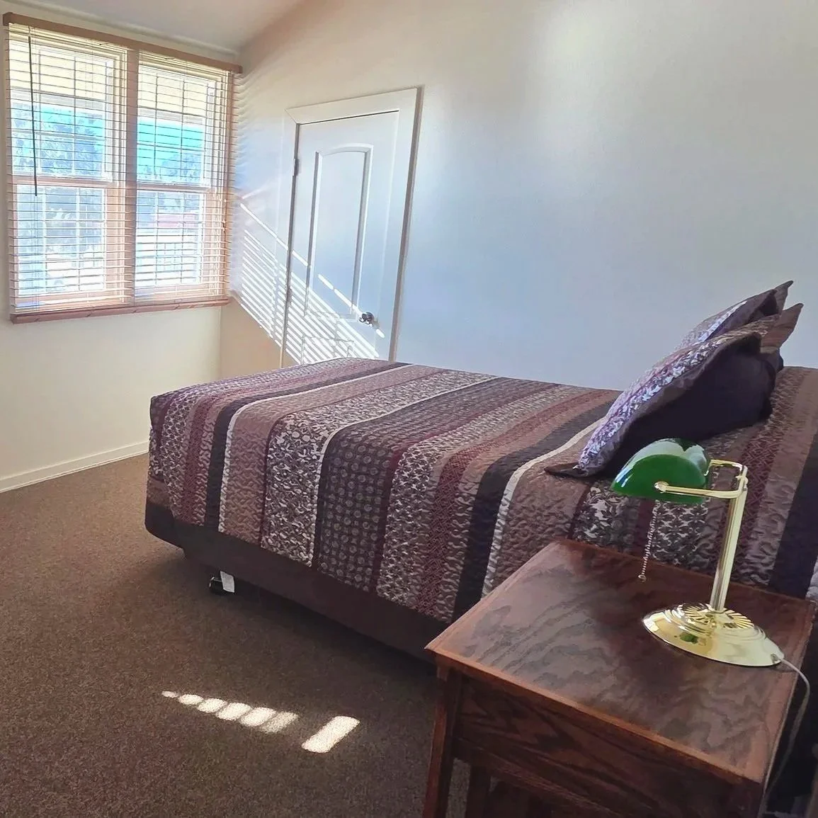 A bedroom with a bed covered in a patterned quilt, two pillows, a wooden nightstand with a green desk lamp, and a window with wooden blinds casting shadows on the wall.