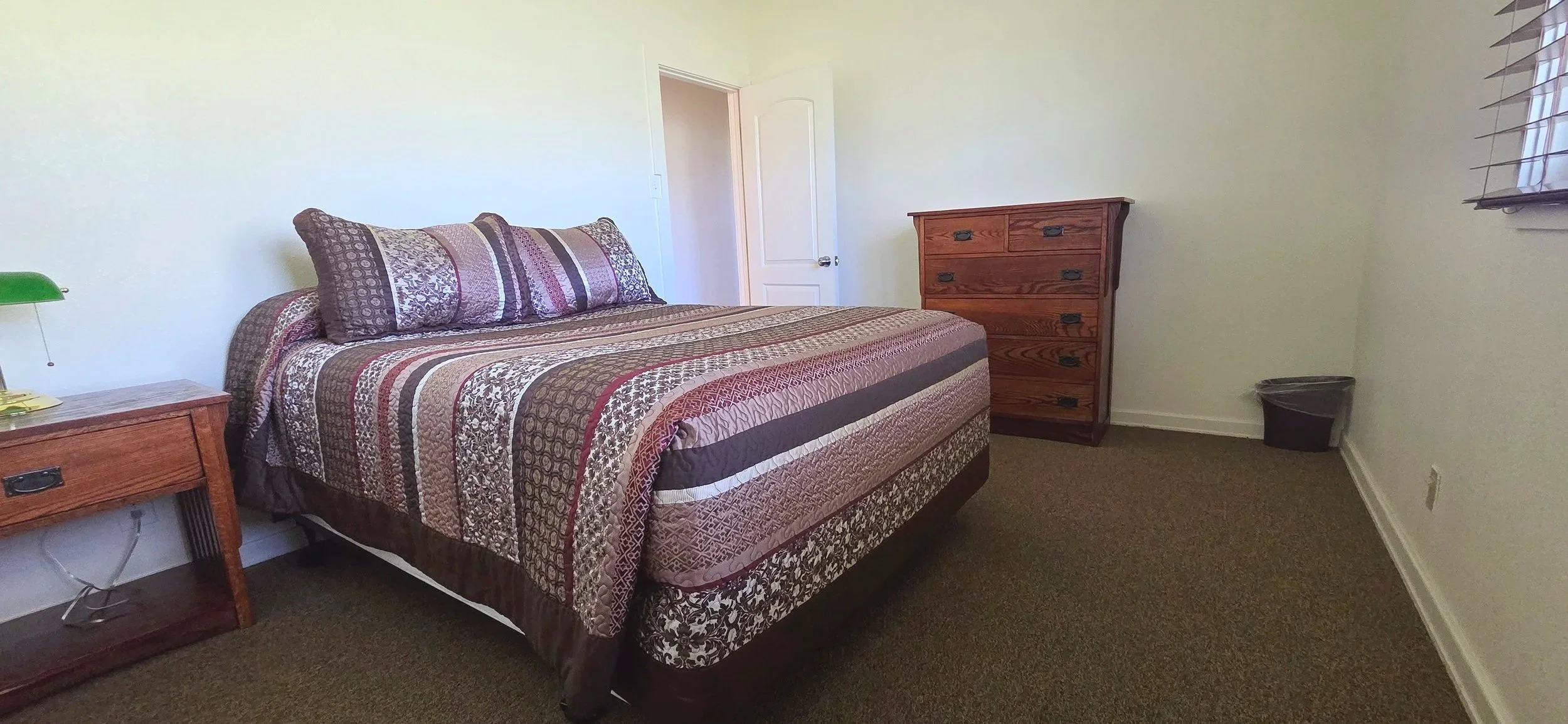 A bedroom with a bed covered in a patterned comforter and pillows, a wooden nightstand with a green desk lamp, a wooden dresser, and a small trash can near the window with blinds.