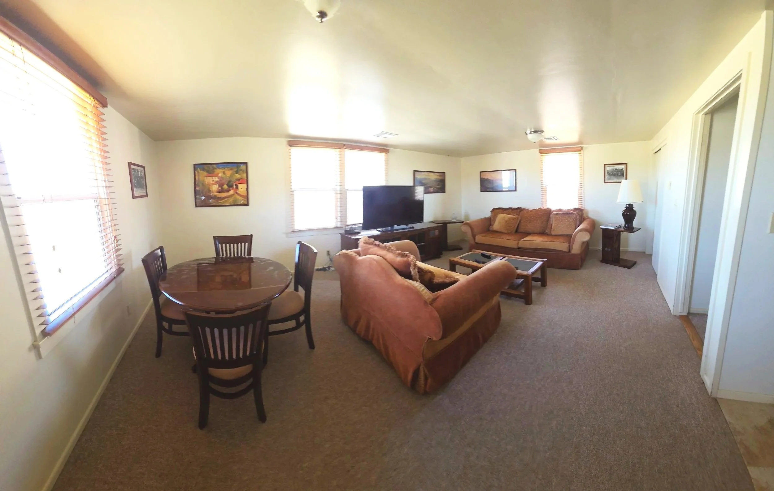 A living room with beige sofas, a flat-screen TV on a stand, a small round dining table with four chairs, and windows with blinds. There are framed pictures on the walls, a lamp on a side table, and a fire extinguisher on the wall near an entrance area with tiled flooring.