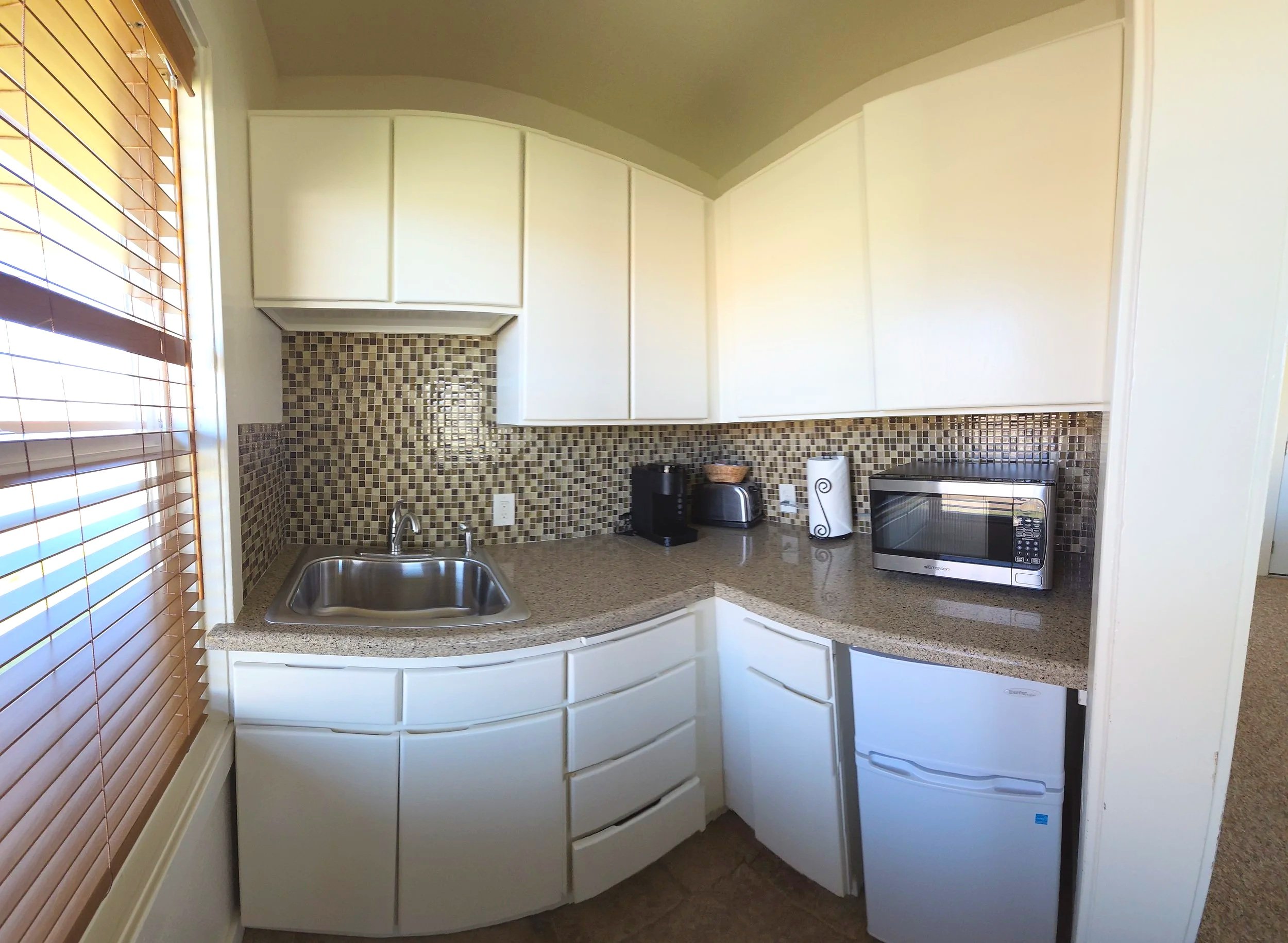 Compact kitchen with white cabinets, granite countertop, mosaic tile backsplash, stainless steel sink, coffee maker, toaster, microwave, small refrigerator, and wooden blinds on window.