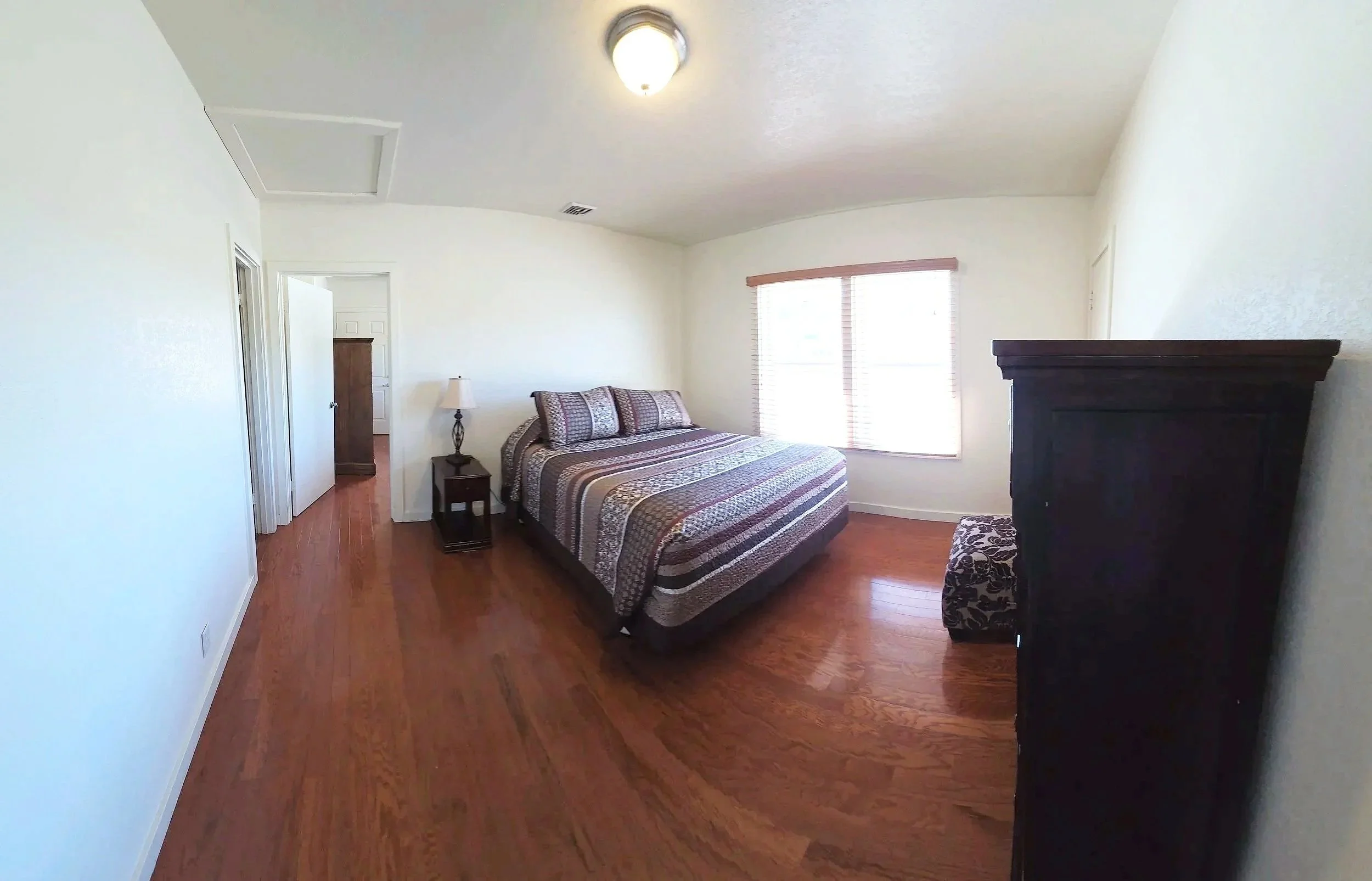 A bedroom with white walls, hardwood floors, a window with blinds, a bed with a patterned quilt, a nightstand with a lamp, and a dark wooden dresser.