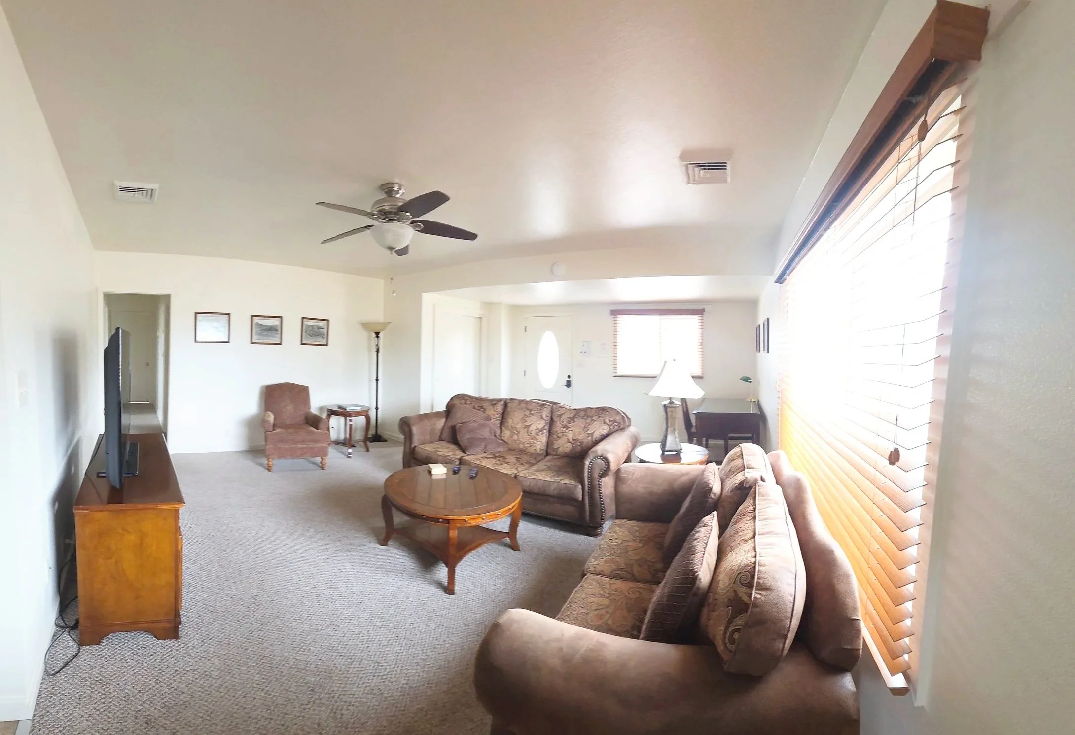 Living room with beige carpet, brown sofas, wooden coffee table, small side table, standing lamp, ceiling fan, large window with wooden blinds, and wall art.