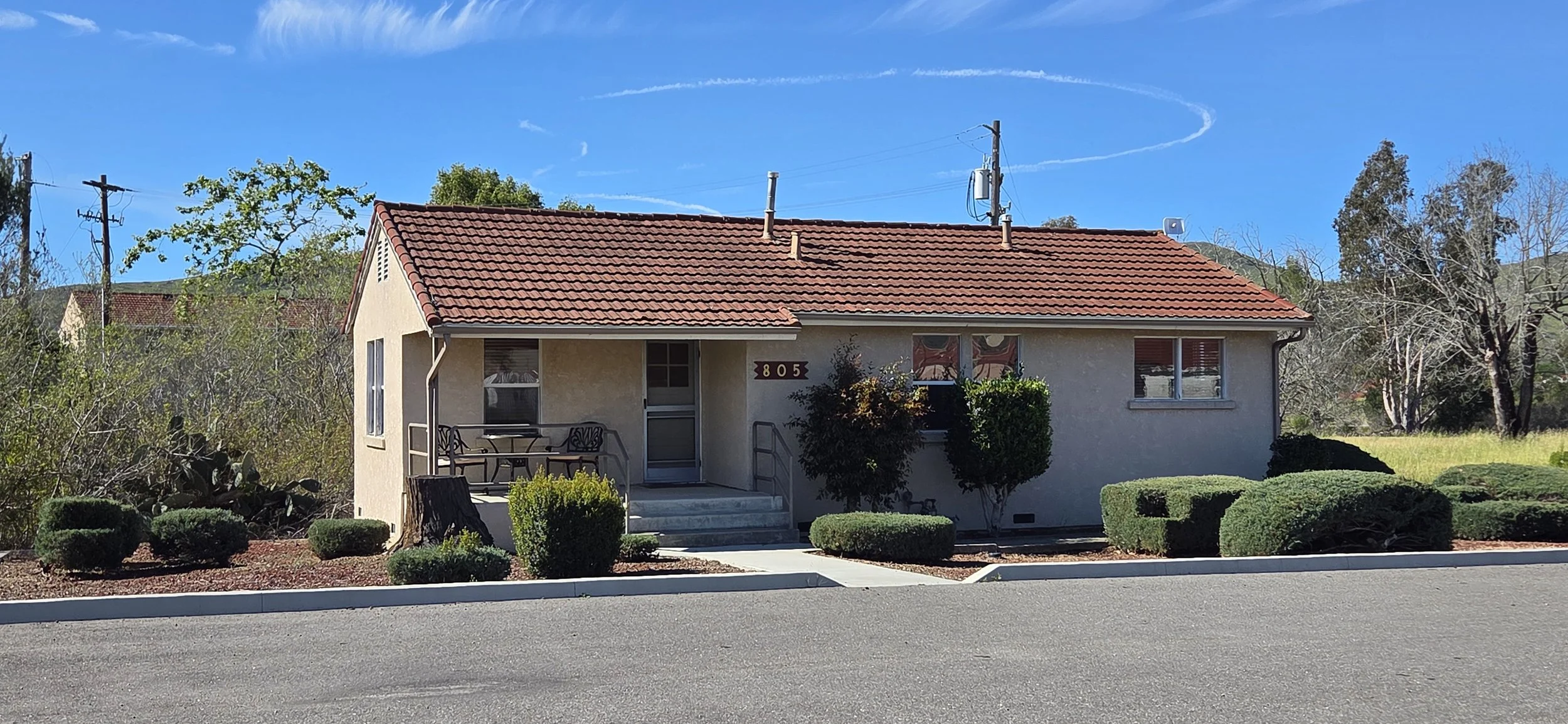 A beige house with a red tiled roof, front porch, and steps leading to the door, surrounded by well-maintained shrubs and trees, with a clear blue sky overhead and a contrail in the sky.