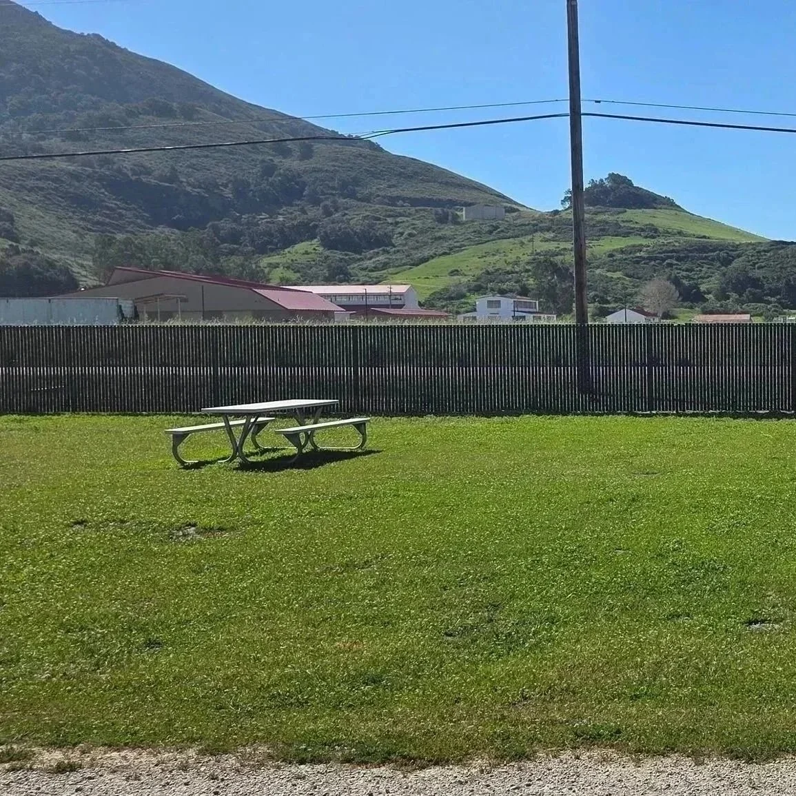 A grassy park with a picnic table and a black fence, with green hills and mountains in the background under a clear blue sky.