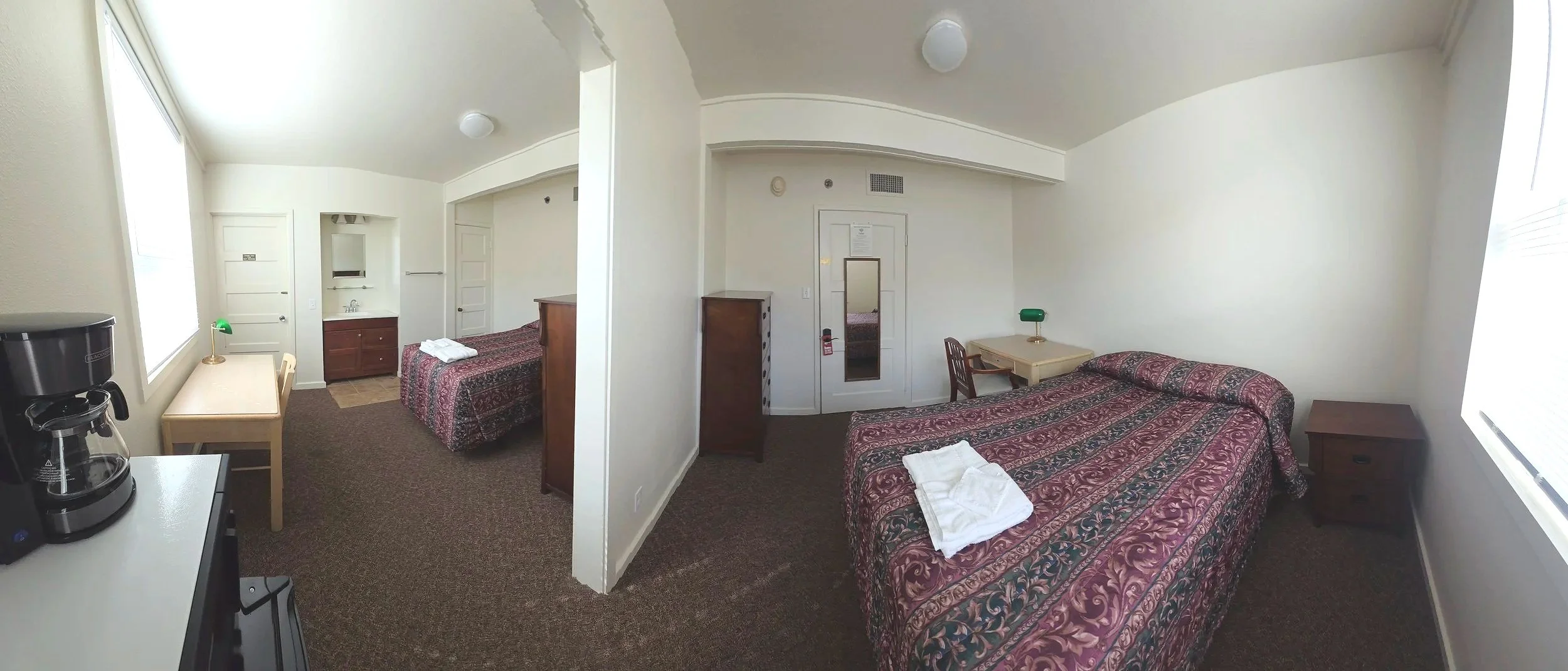 Inside a motel room with two beds, nightstands, a small table with a green lamp, a mirror, a door, a window with blinds, and a coffee maker on a counter.