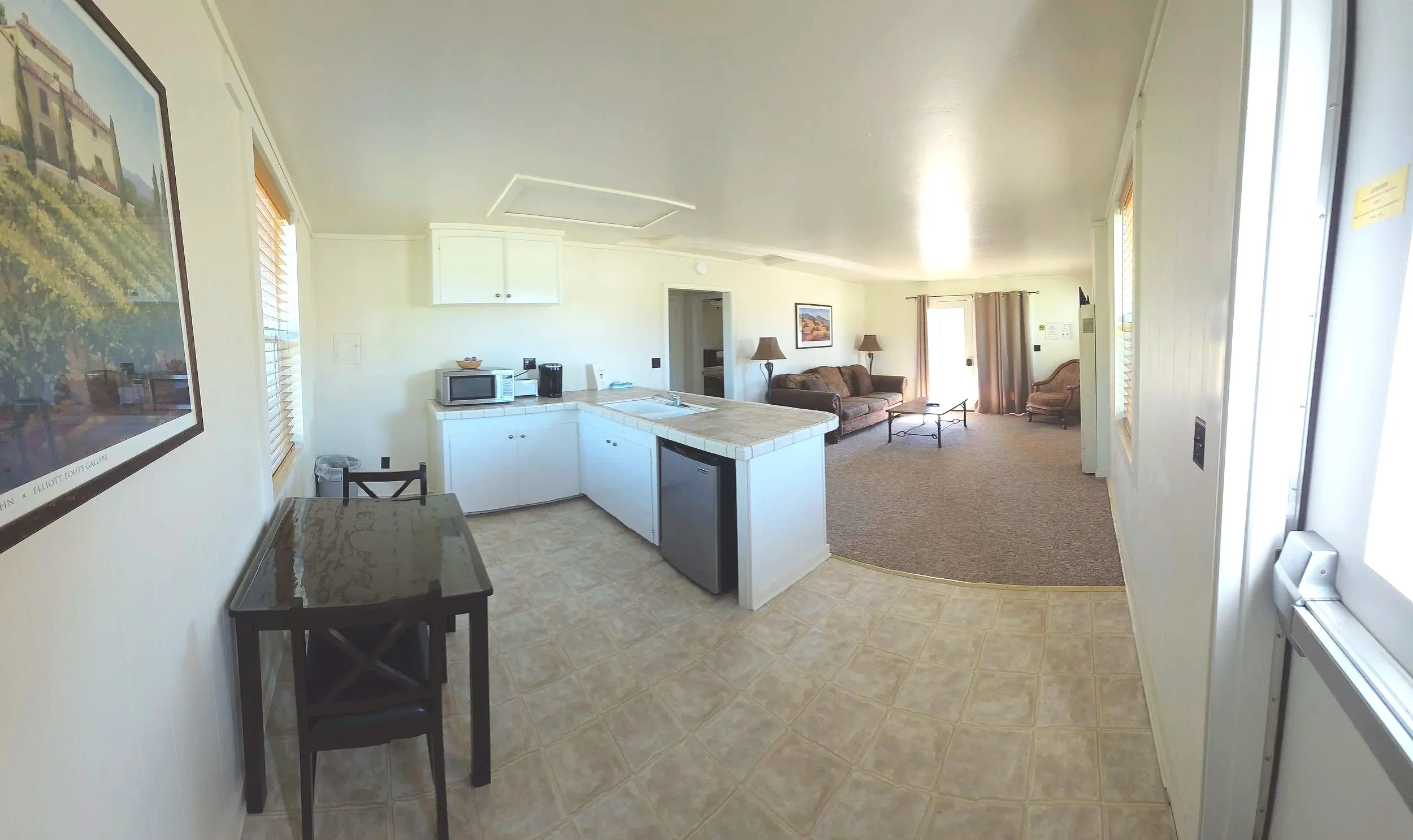 Open-concept living area with kitchen, dining, and living room, showing a tiled kitchen area with microwave, refrigerator, and a small dining table, expanding into a carpeted living room with sofas, coffee table, lamps, and a patio door with curtains.