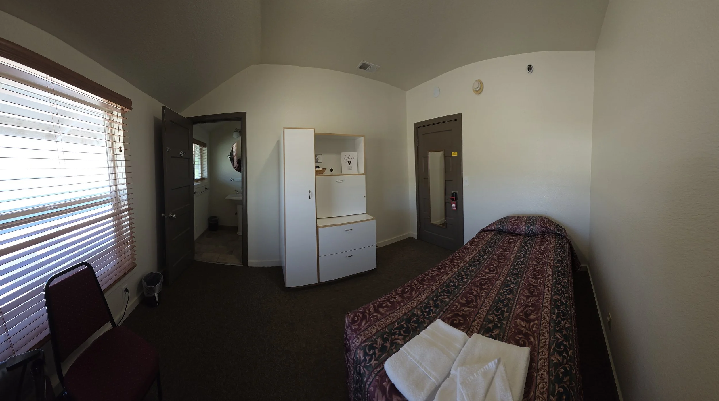 Small hotel room with single bed, window with blinds, white cabinet, dark door, and bathroom visible through open door.